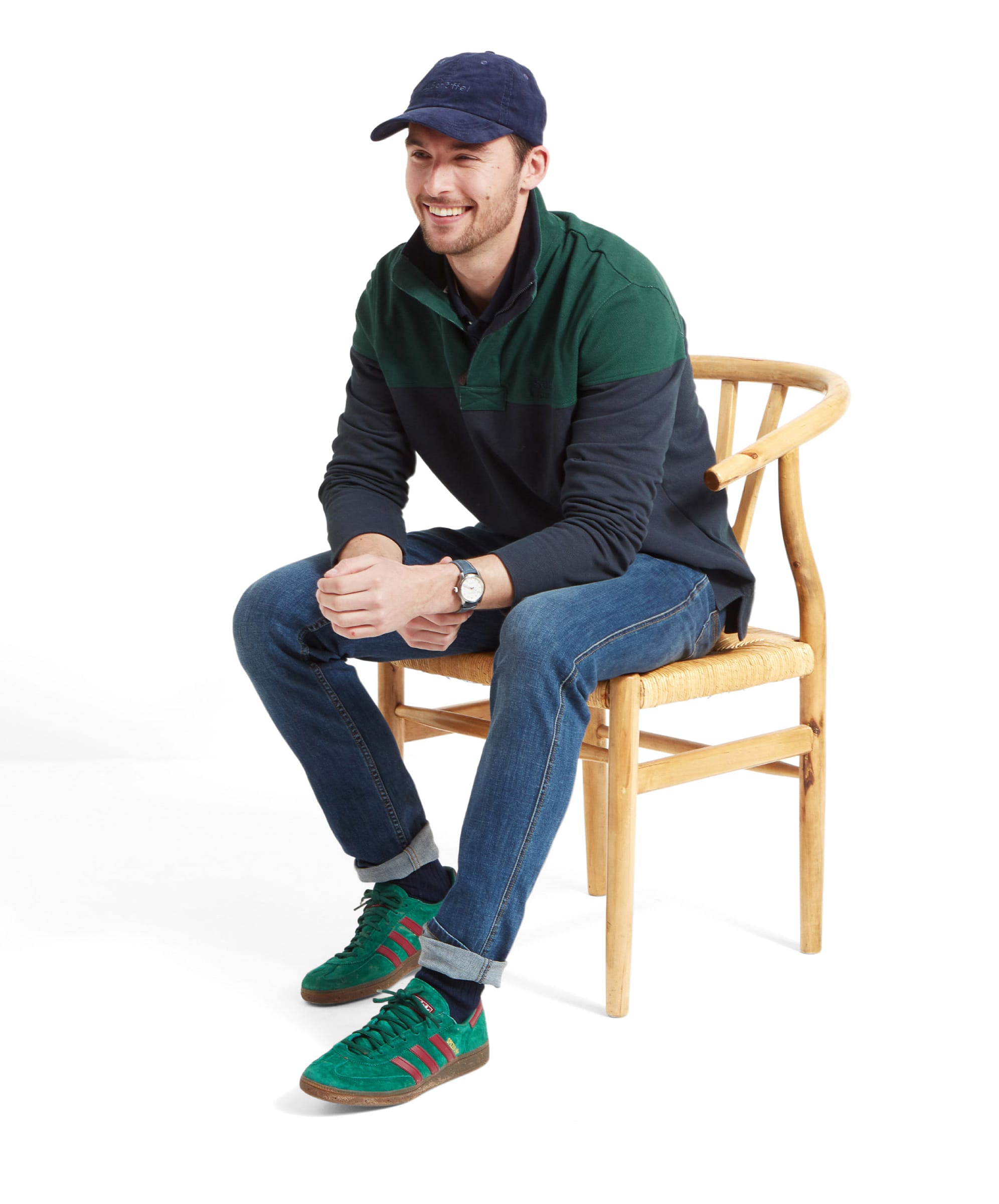 A man sits of a wooden chair  whilst wearing the Schöffel Helford Heritage Sweatshirt for Men in Green with a blue cap, navy jeans and green trainers. He's looking to his left whilst smiling.