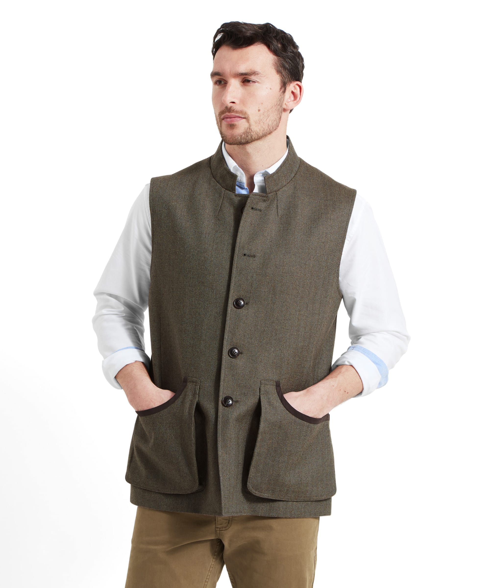 A man wears the Schöffel Holcot Tweed Waistcoat for Men in Green with a white shirt with the sleeves rolled up his forearms. He has both hands in his pockets and looking to his left.