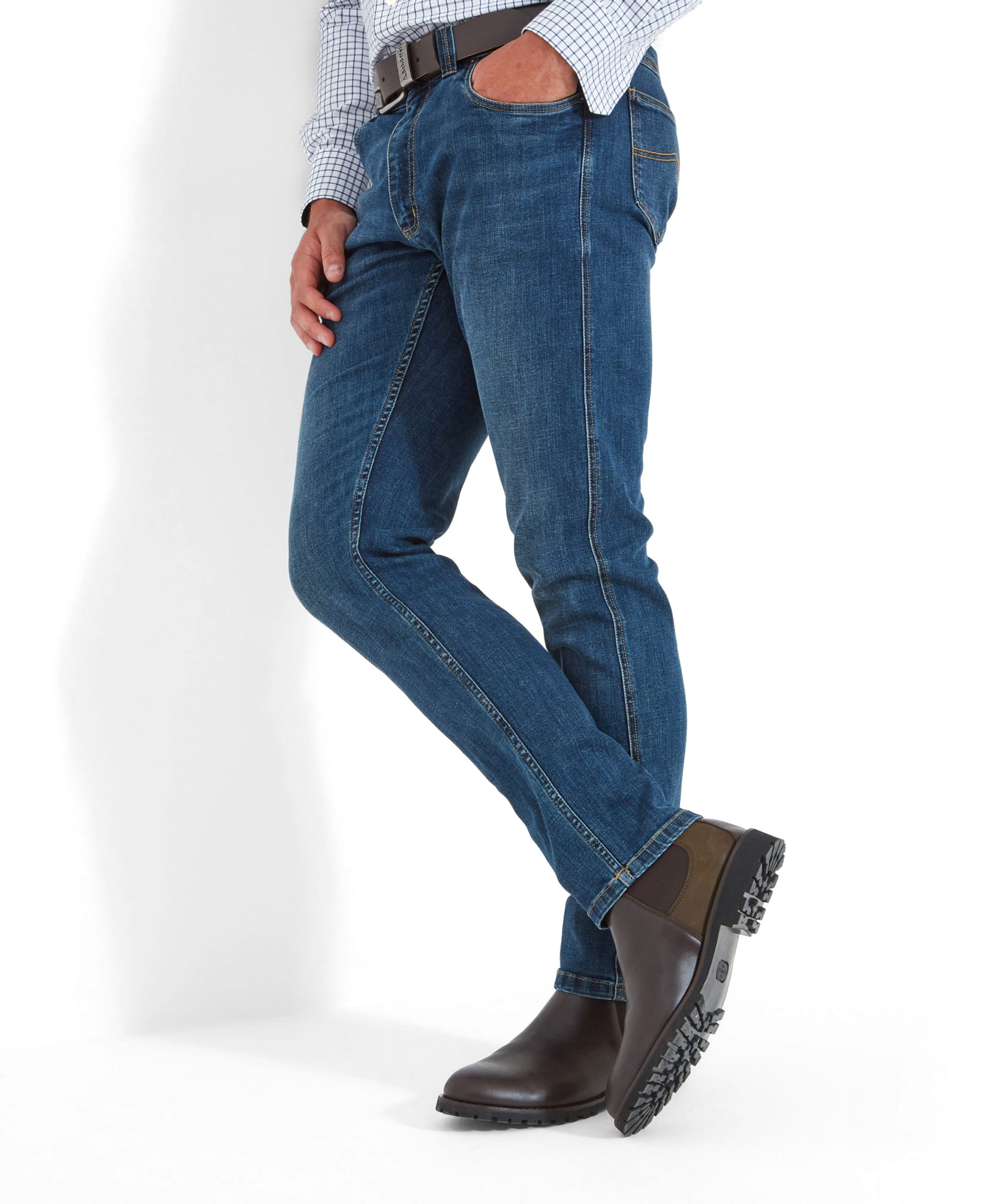 A man wears the Schöffel James Jean for Men in Blue with brown boots and a dark brown leather belt. The model appears to be leaning and has their right hand in the jean pocket.