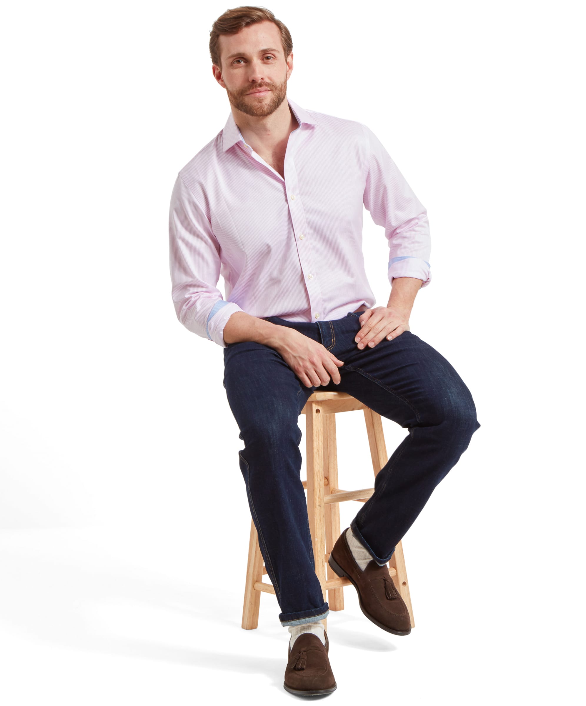 A model sits on a wooden stool and rests his hands in his lap whilst wearing the Schöffel James Jean for Men in Navy, a pink stripe shirt, and brown suede shoes.