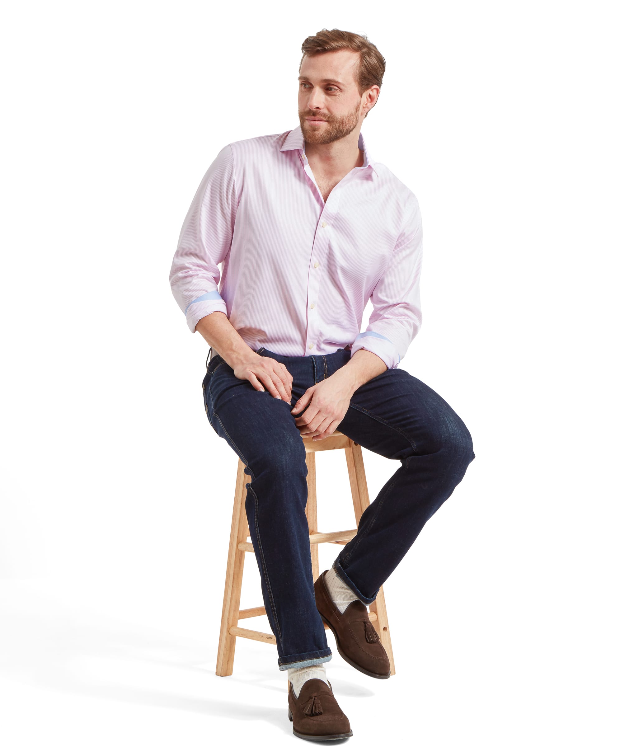 A model sits on a wooden stool and rests his hands in his lap whilst looking away to the left. He's wearing the Schöffel James Jean for Men in Navy, a pink stripe shirt, and brown suede shoes.