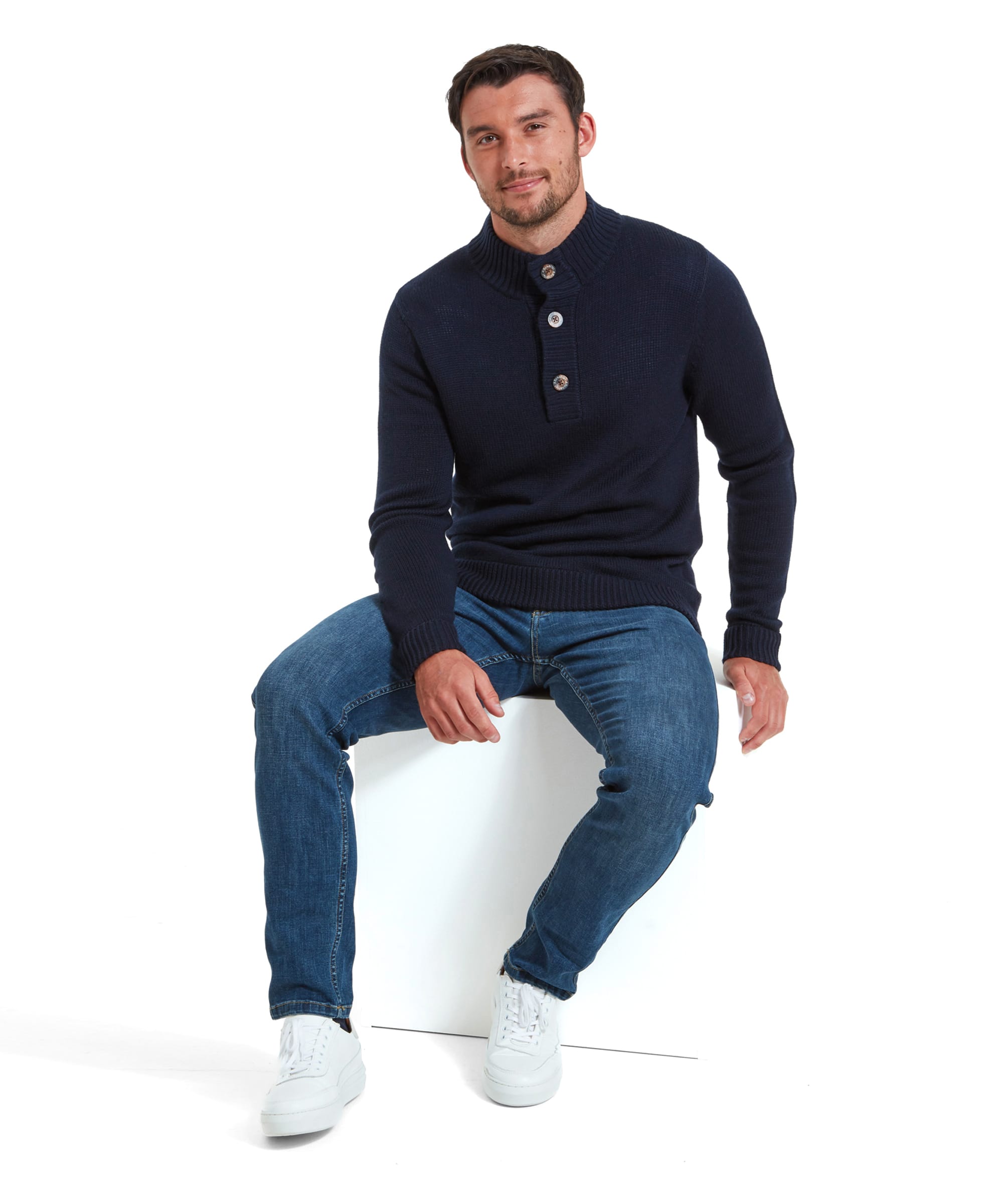 A man wears the Schöffel Kenmore Lambswool Mix Jumper for Men in Navy with denim jeans and white trainers. The model is seen sitting.