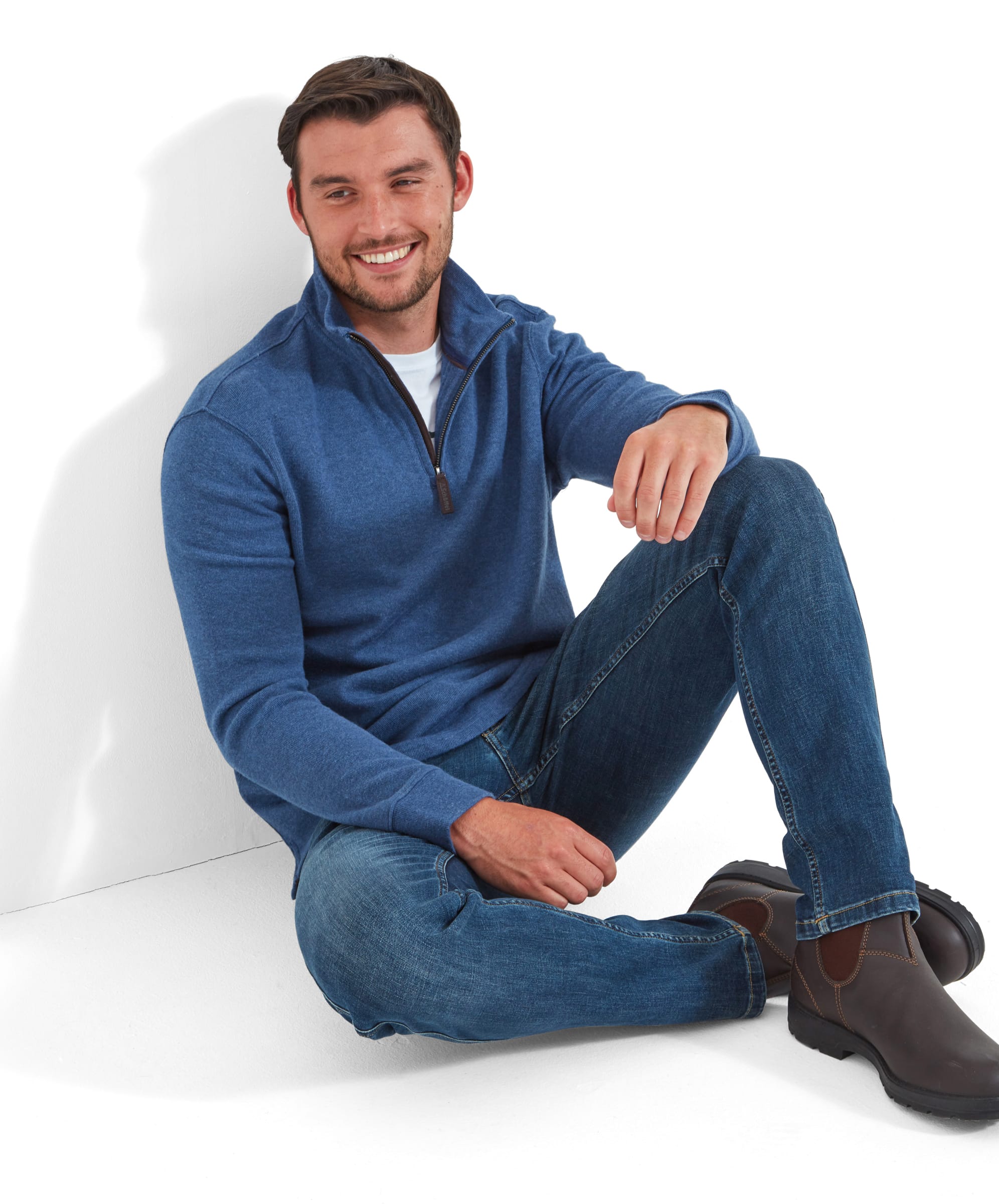 A man in seen sitting on the floor, leaning on a wall wearing the Schöffel Kingsbridge Cotton French Rib 1/4 Zip for Men in Blue, partnered with a white t-shirt, denim jeans and dark brown boots.