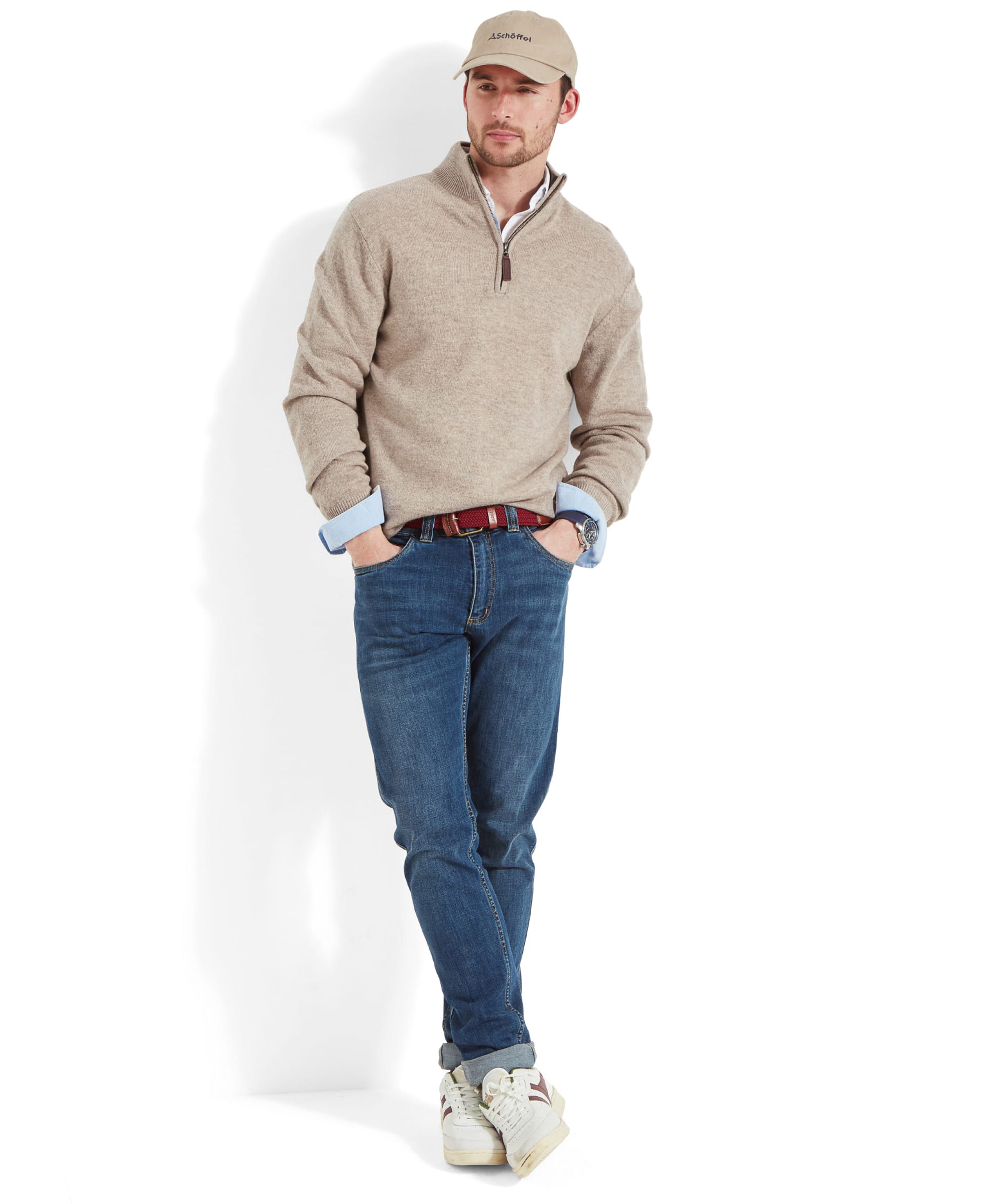 Full body view of the Schöffel Lambswool 1/4 Zip for Men in Light Brown as a model leans against a background, with his hands placed in his jean pockets. His outfit is made up of a beige cap, a white shirt underneath a sweater, red woven belts, denim jeans, and white trainers.