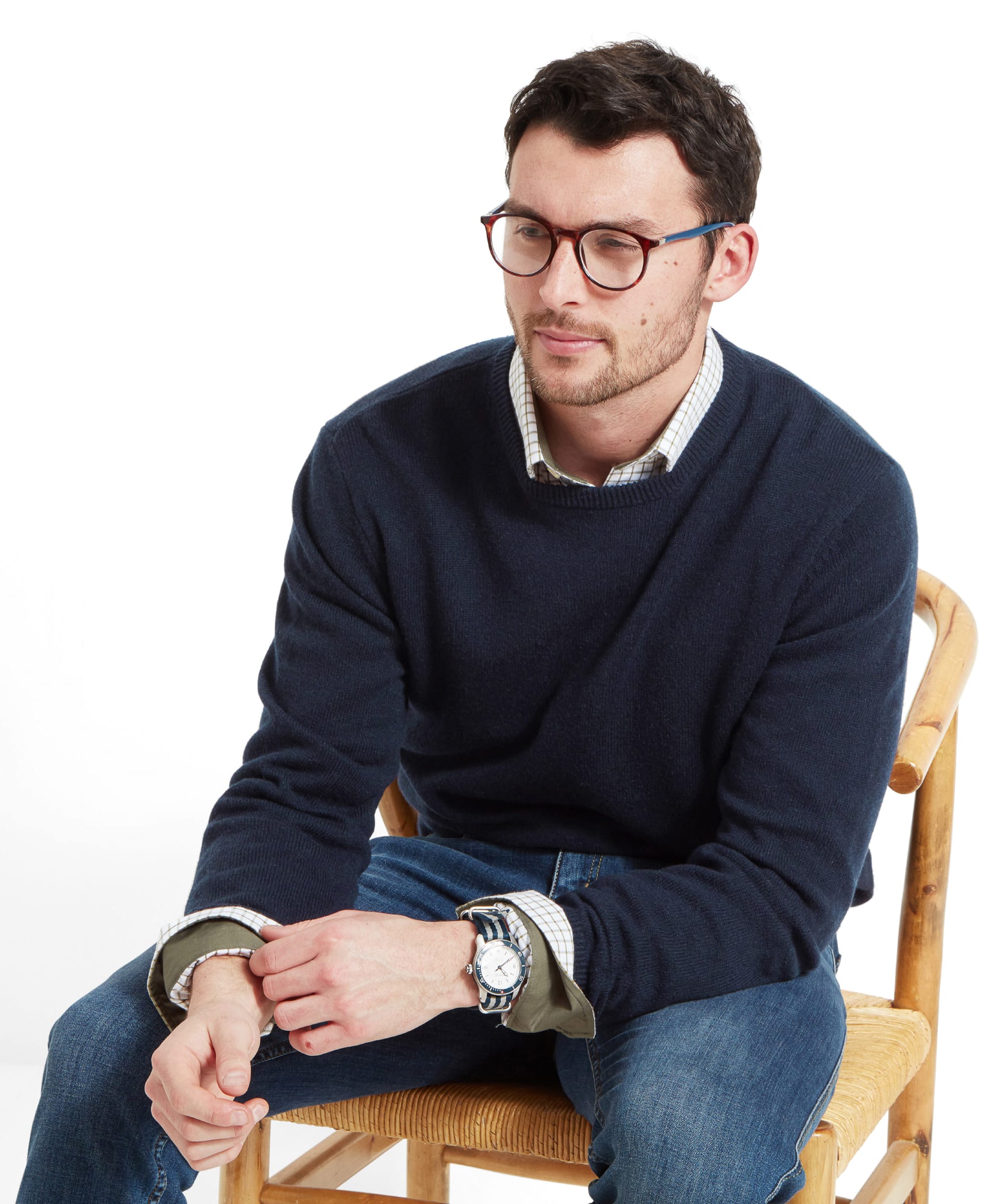 A man wears a Schöffel Lambswool 1/4 Zip for Men in Navy with a check shirt, Navy watch, and denim jeans whilst sitting on a wooden chair.