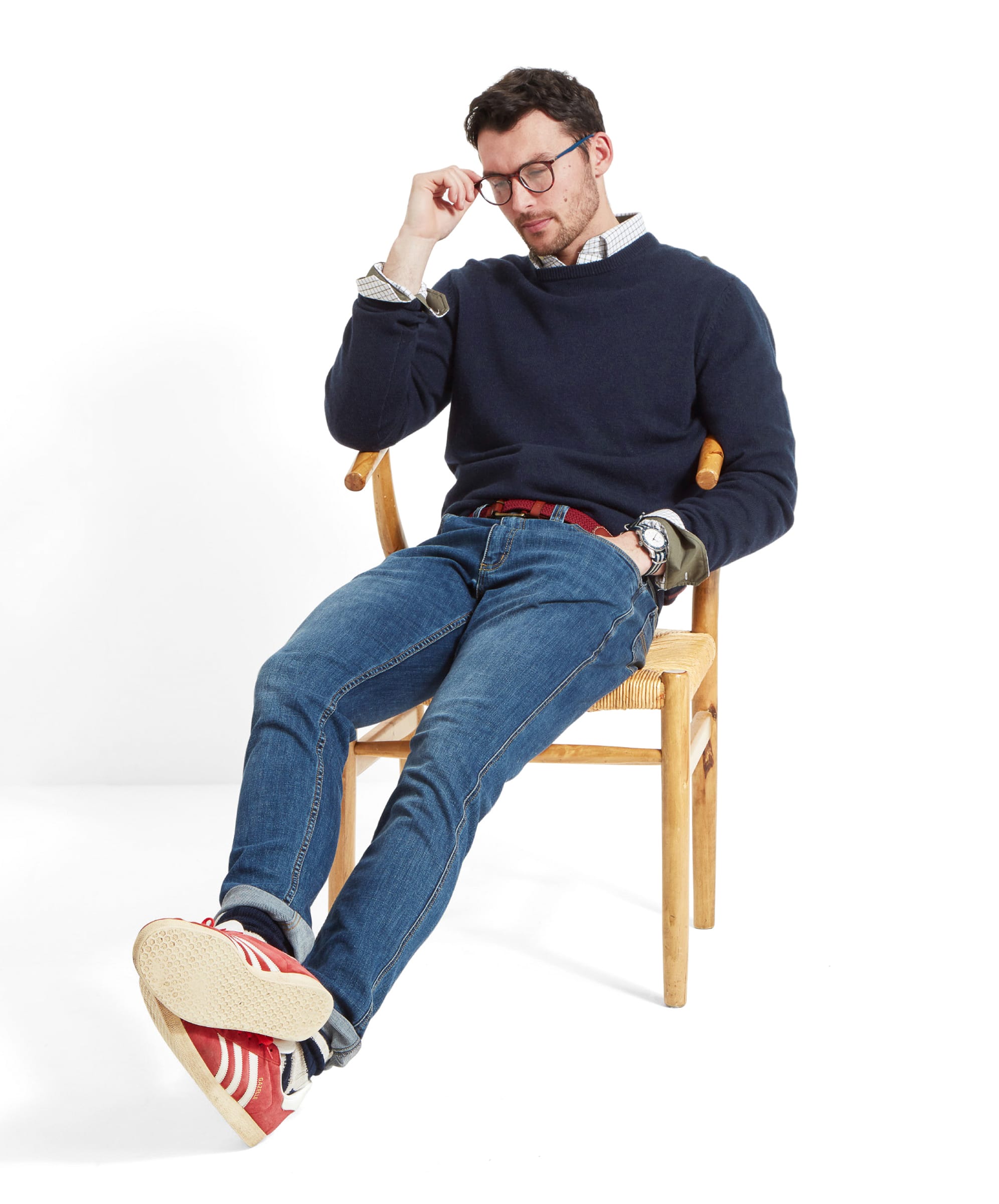 Full body shot of a man relaxing on a wooden chair adjusting his glasses. He wears the Schöffel Lambswool 1/4 Zip for Men in Navy with a check shirt, red woven belt, denim jeans and red trainers.