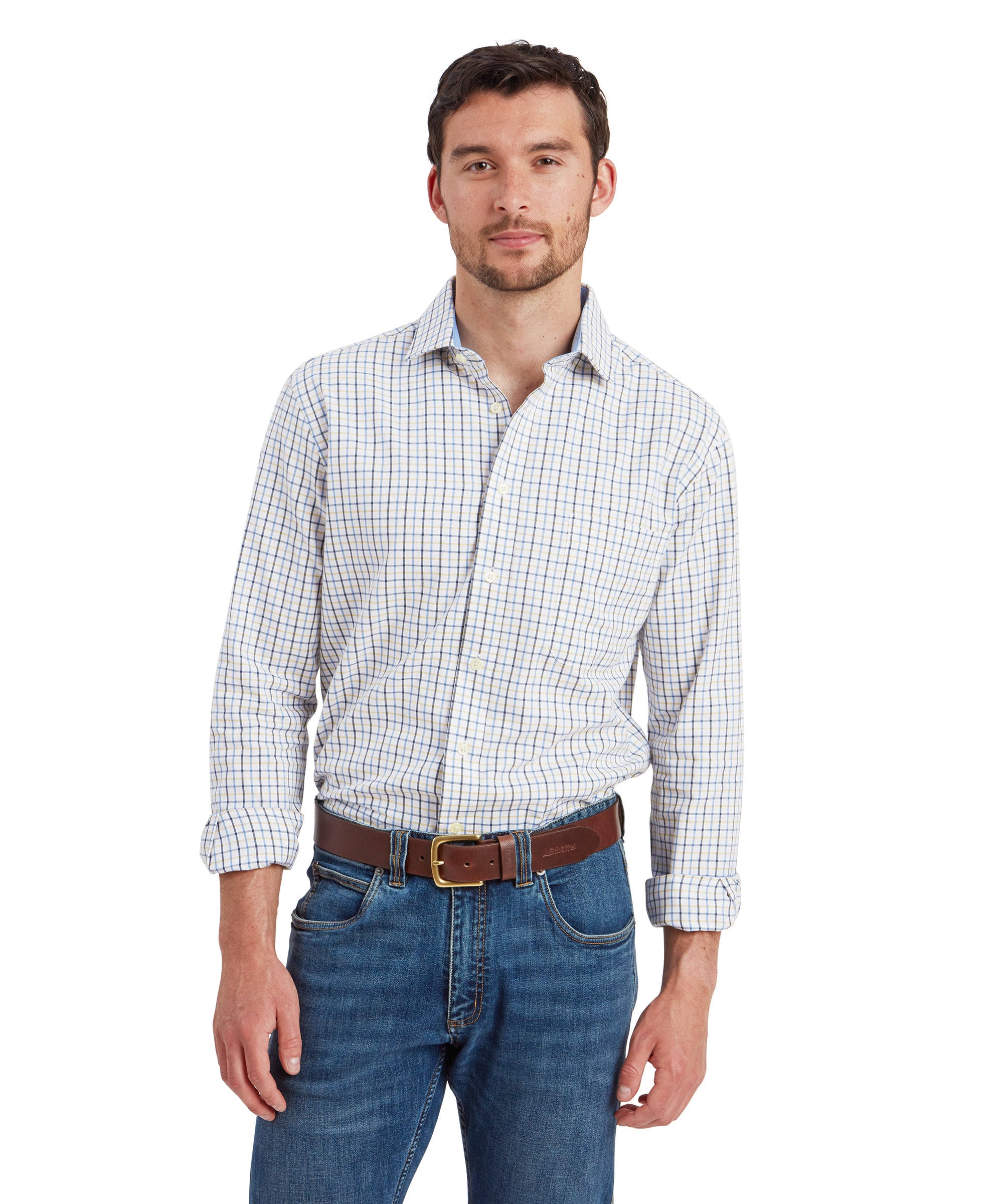 A man wearing a long-sleeved, Schöffel Milton Tailored Shirt for Men in Blue with a pattern of blue, yellow, and white checks. He is standing with his arms relaxed at his sides, smiling slightly. The shirt is tucked into a pair of blue jeans, and he is wearing a brown leather belt. The shirt features a button-down collar and a pocket on the left side of the chest.