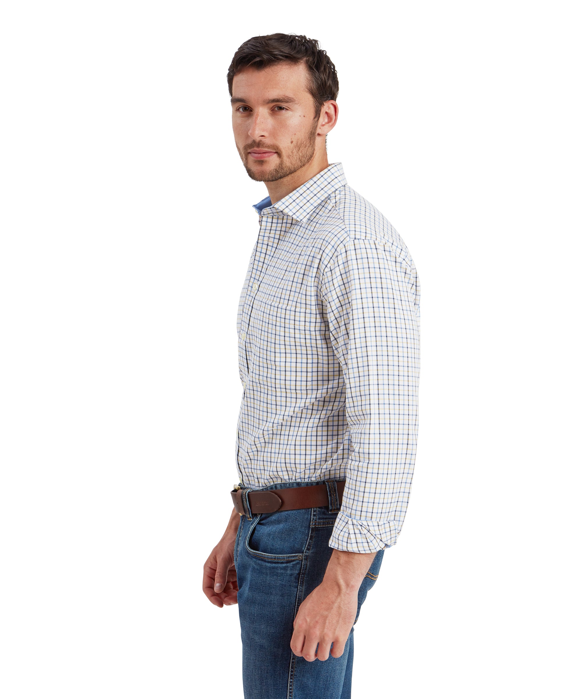 A side profile view of the man in the Schöffel Milton Tailored Shirt for Men in Blue and blue jeans. He is standing upright, looking slightly over his left shoulder. The shirt shows a well-fitted silhouette, with the shirt's checks visible along the sleeve and the side of the body.