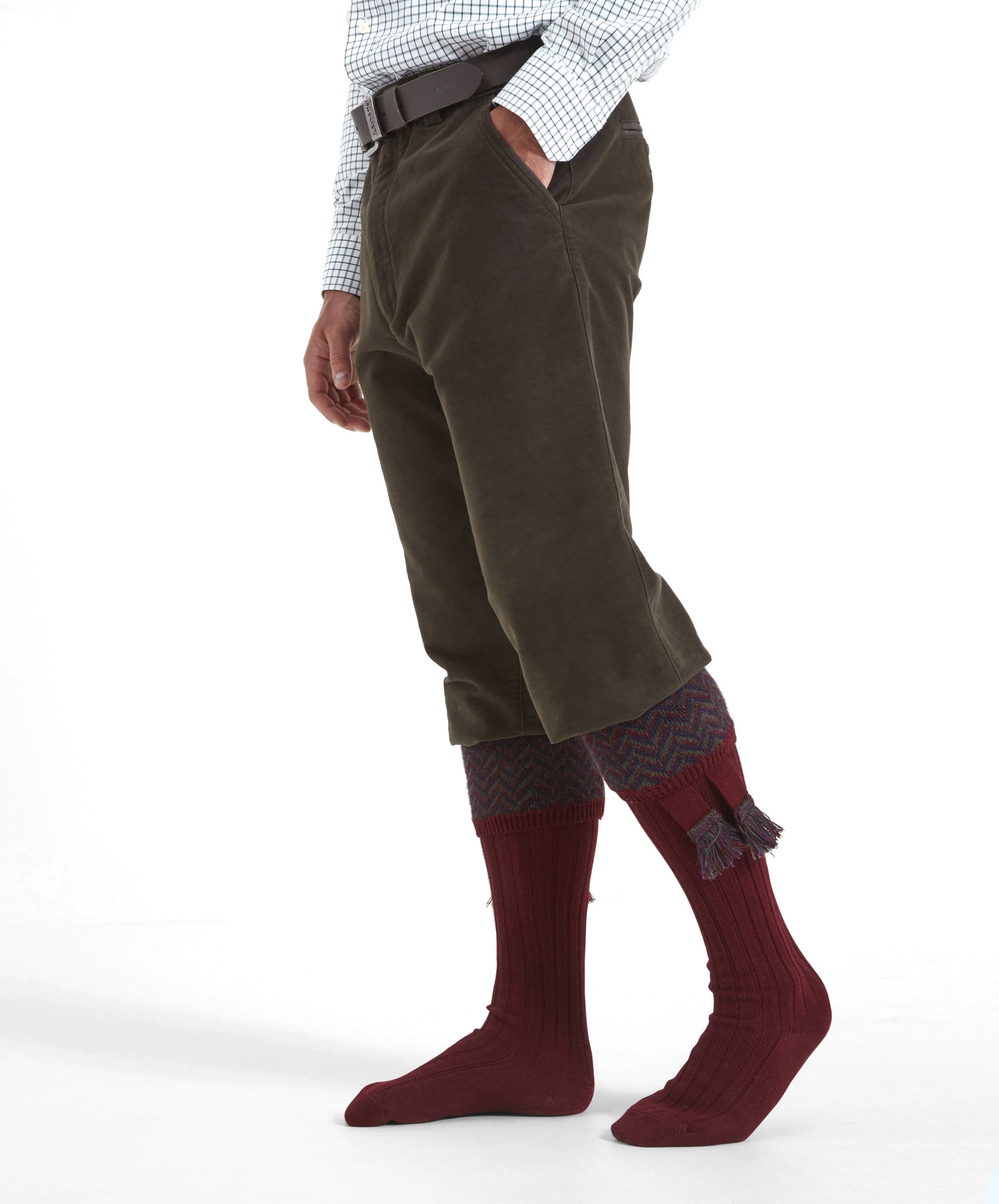 A side view of the Schöffel Moleskin Trousers for Men in Green as a man holds his hand in the trouser pocket. The fit of the trousers is loose and similar to that of a breek. The fabric has a soft appearance and are worn with tall burgundy red tassle shooting socks.