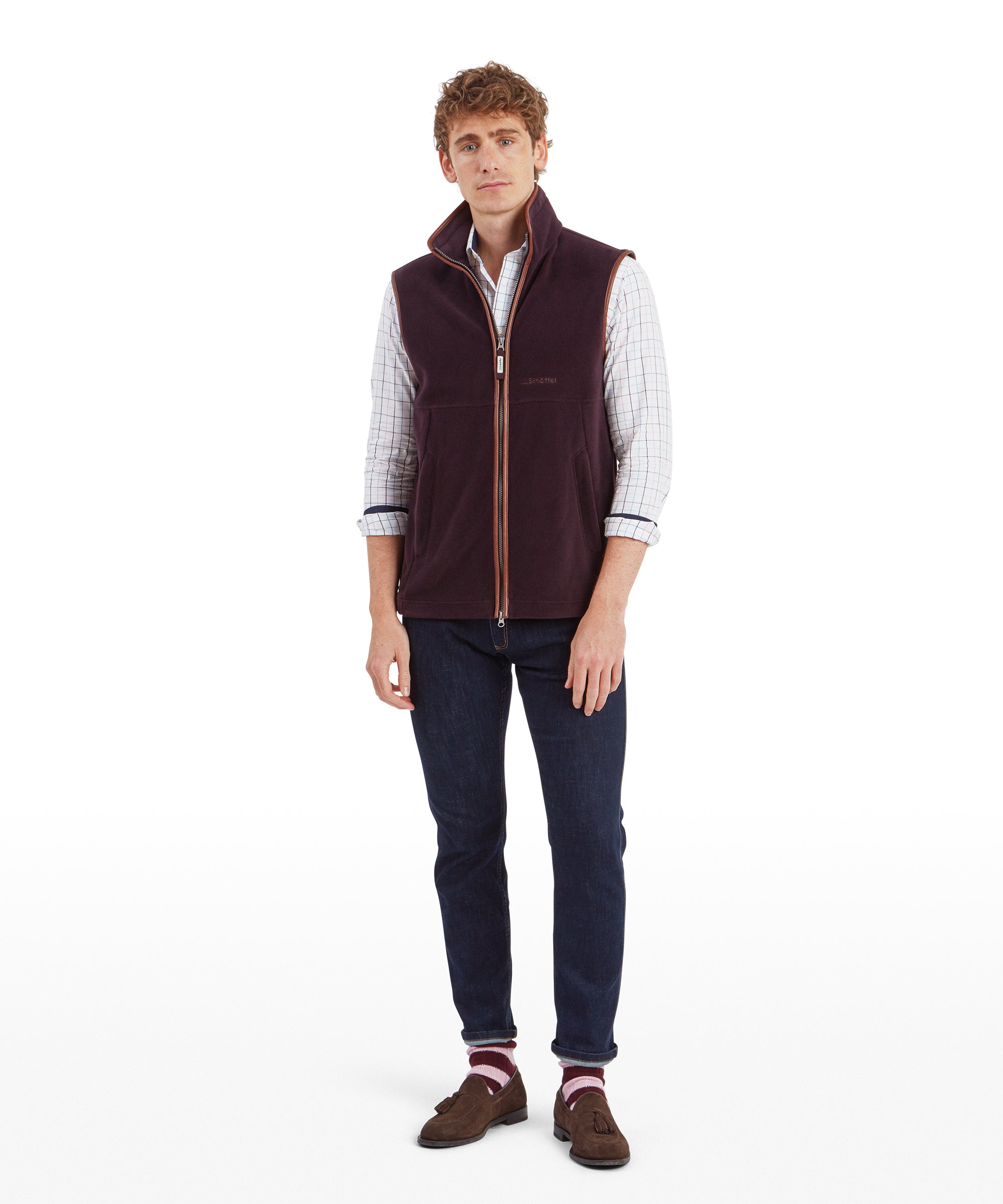 A full-body view of the same man modelling the Schöffel Oakham Fleece Gilet for Men in Brown with a light blue and purple check shirt, dark denim jeans with the hems folded up showing some eccentric pink and burgundy stripe socks with dark brown suedette brogues. The image highlights the decent length to the gilet and the loose fit.