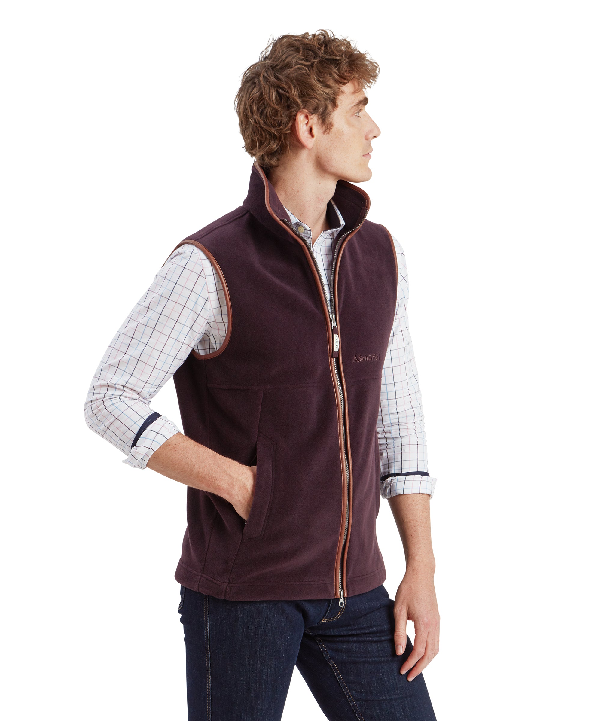 Side view a male with mousey brunette curly hair stands looking away wearing the Schöffel Oakham Fleece Gilet for Men in Brown whilke resting a hand in the gilet pocket. The man partners the gilet with a light blue and purple long sleeve button-down shirt with the cuffs rolled up his forearms. The collar on the gilet is tall and sits almost to the edge of his jaw.