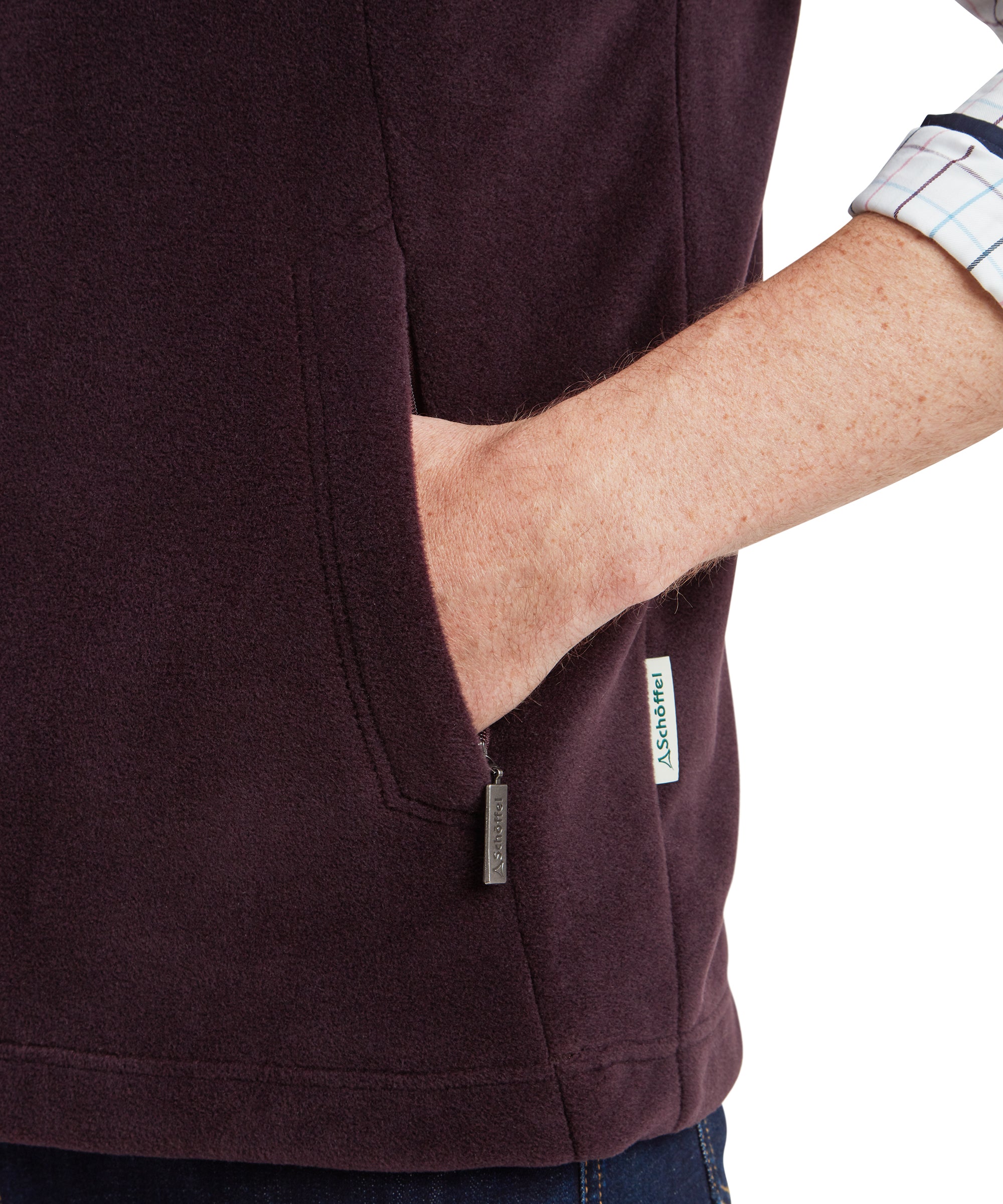 Close-up on the front pocket on the Schöffel Oakham Fleece Gilet for Men in Brown highlighting the double stitching design and the metal zip pull on the pocket zipper. The man holds his hand in the pocket to show the generous size. Down the side seam a Schöffel branded pip can be seen.