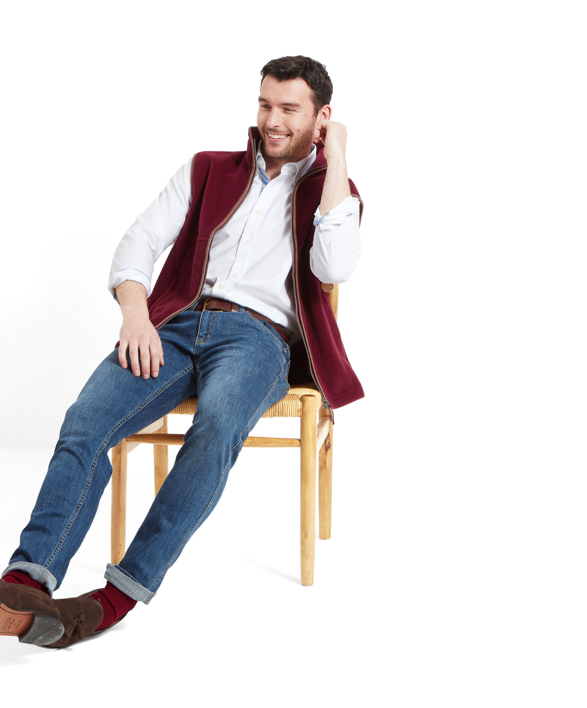 The model is seated on a wooden chair, smiling and looking relaxed while wearing the Schöffel Oakham Fleece Gilet for Men in Red.