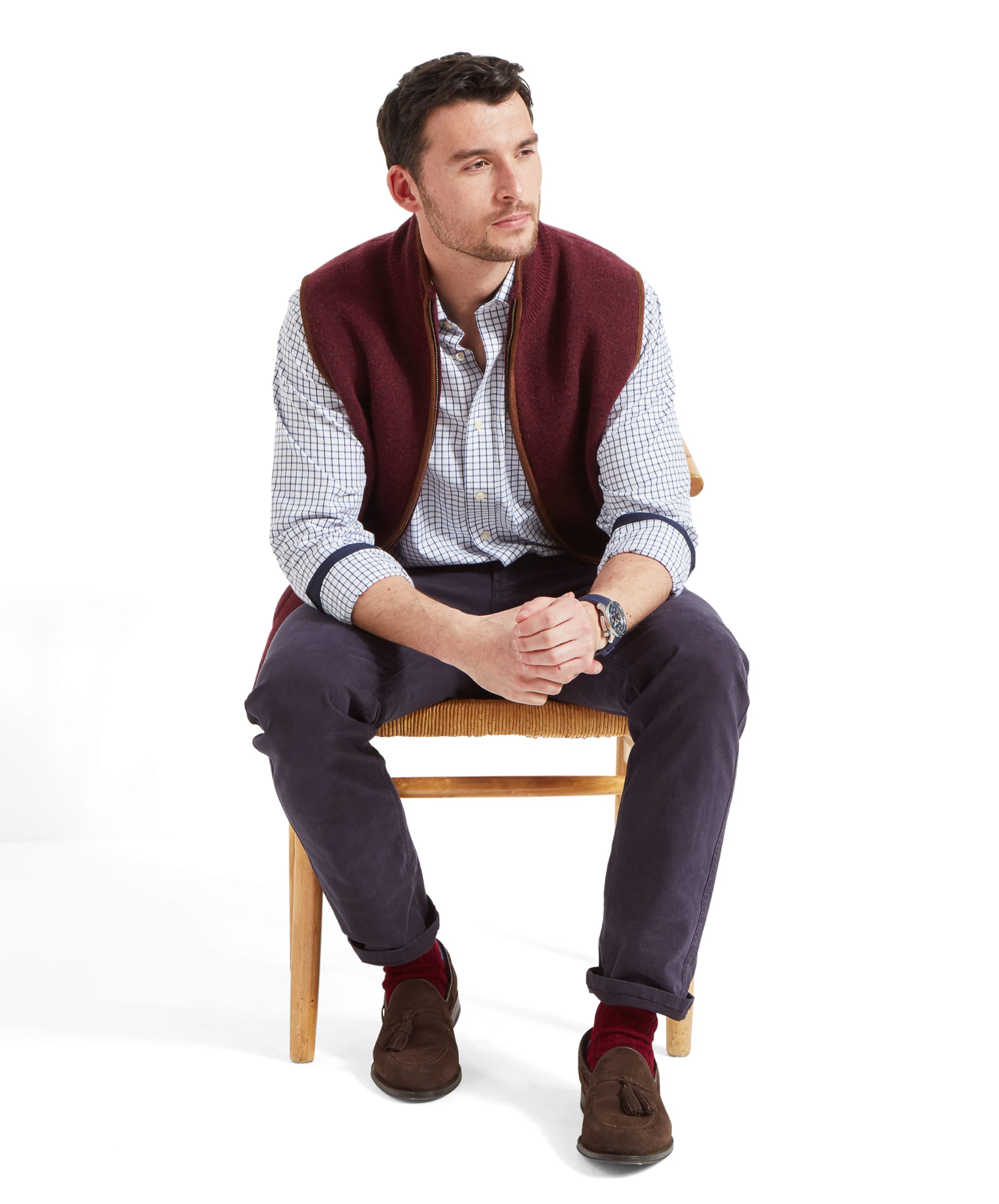 Seated model showcasing the Schöffel Orkney Merino Gilet for Men in Red, paired with a checkered shirt and dark trousers.