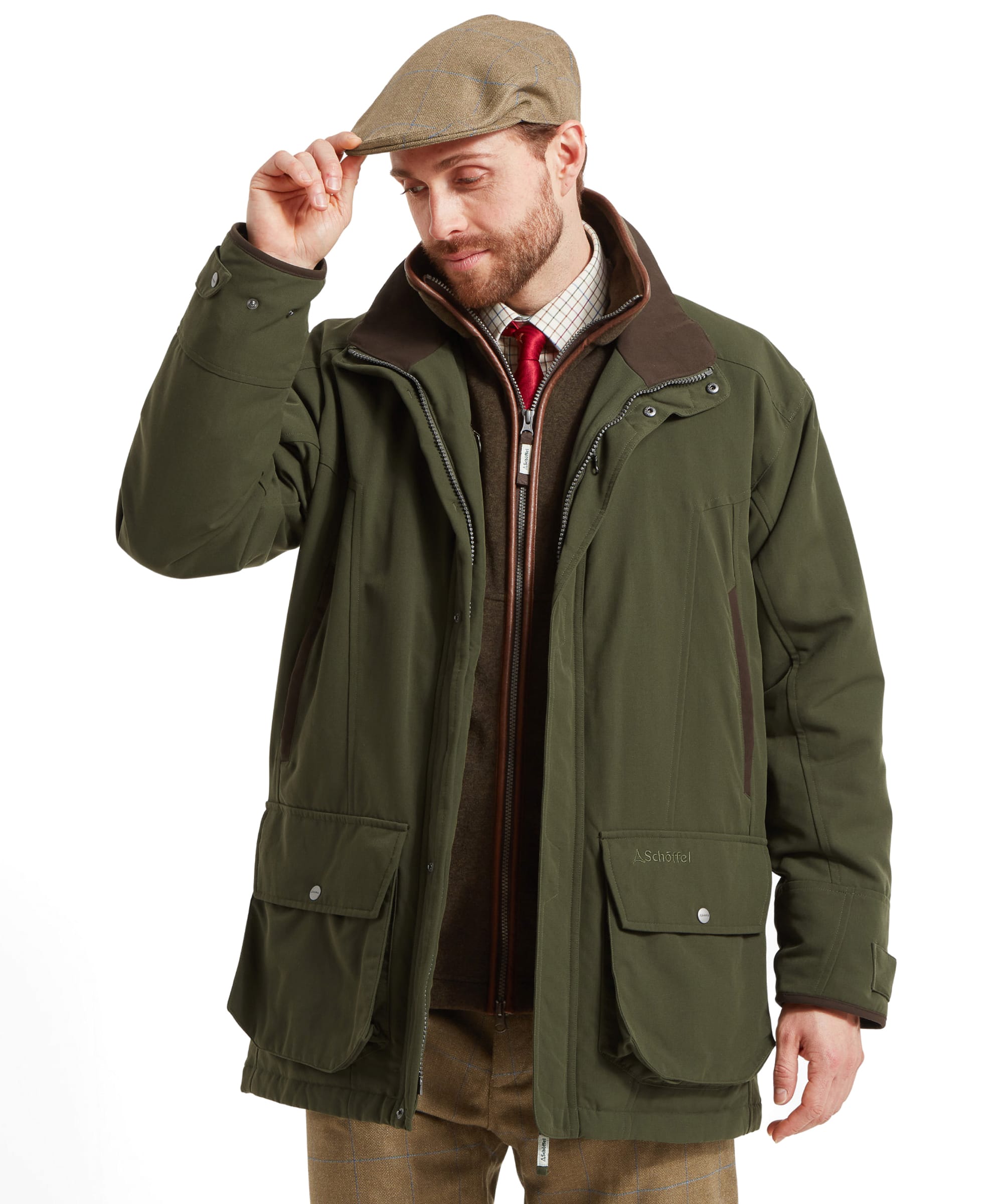 A man in the Schöffel Ptarmigan Classic Coat for Men in Green, touching the brim of his hat and looking down, showcasing the coat's details and the layered outfit underneath.