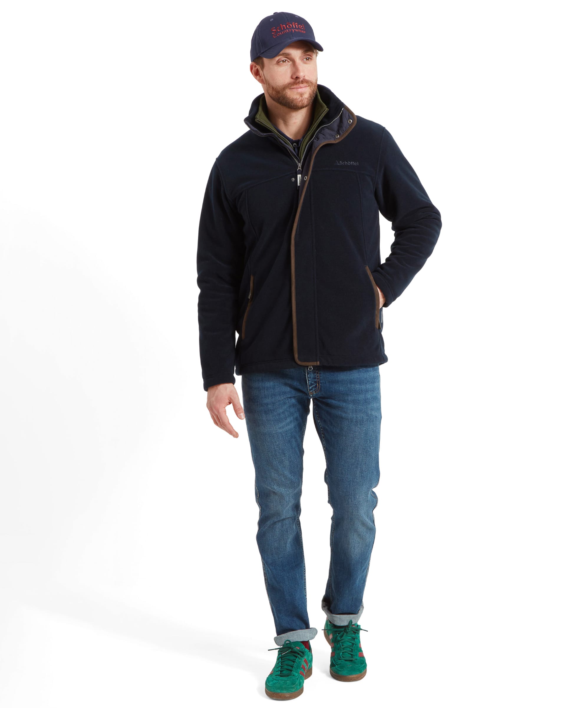 Full body profile of a man modelling the Schöffel Rutland Fleece for Men in Navy. He stands with his right hand in his pocket and is peering to the side. He wears the jacket with a navy blue cap, a green sweater underneath , denim jeans and green trainers.