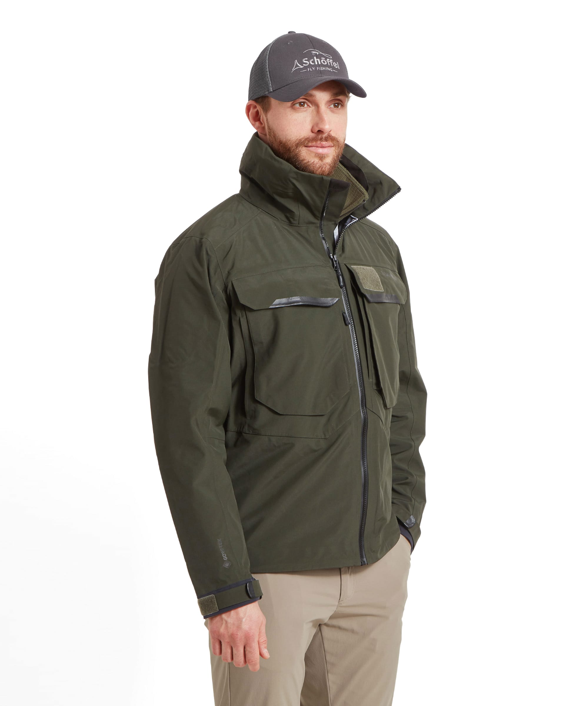 A side three-quarter view of a man wearing the Schöffel Salar II Wading Jacket for Men in Green zipped up with a hand in the pocket of his trousers. He wears a grey cap and beige trousers whilst looking .into the distance.