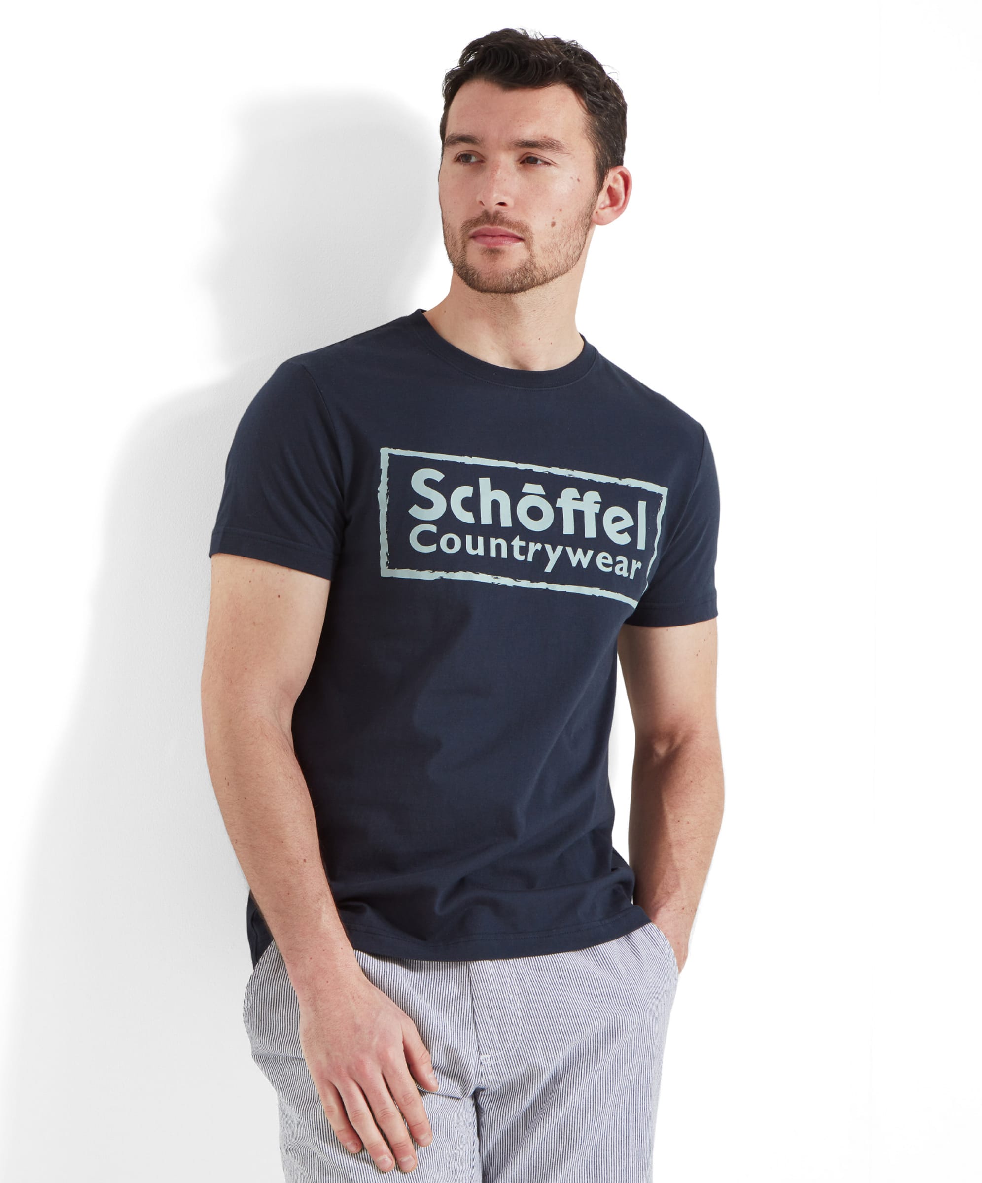 A man wears the Schöffel Heritage T-Shirt for Men in Navy with white and blue stripped shorts. The model is casually leaning against the back drop and holds his right hand in his pocket.