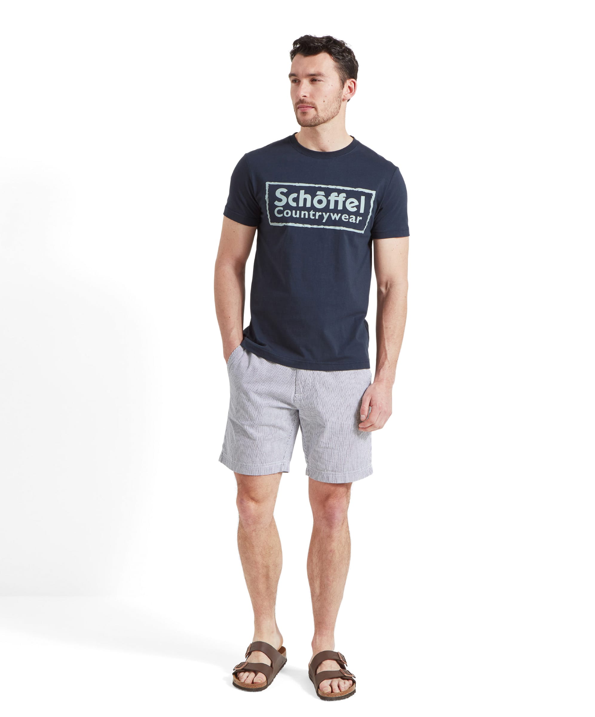 Full body shot of a man modelling the Schöffel Heritage T-Shirt for Men in Navy with white and blue horizontal stripped shorts and brown sandals showing a summery outfit.