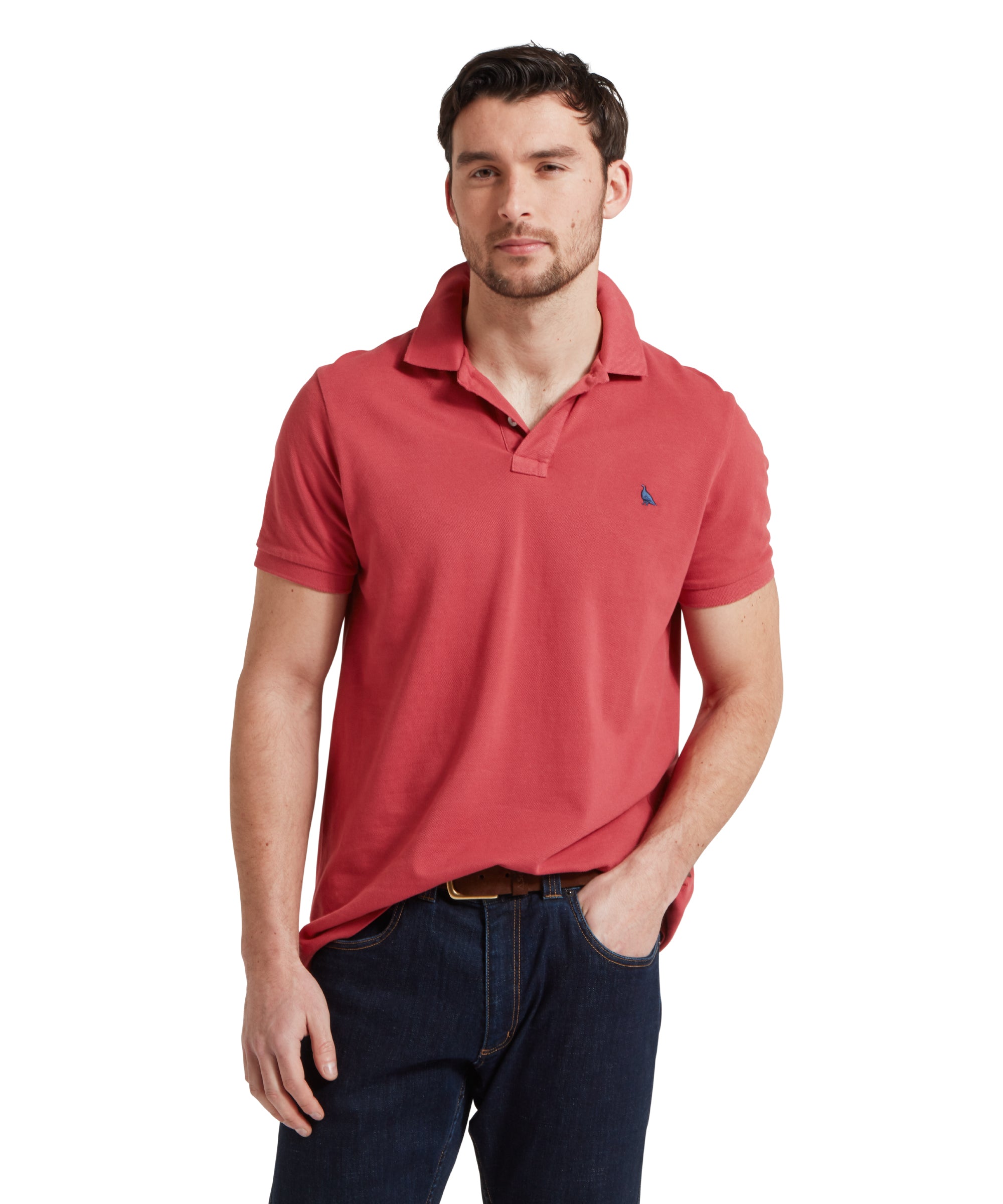 A close up image of a male model wearing a red Schöffel polo shirt with a small embroidered bird logo on the chest, paired with dark denim jeans and a tan leather belt. He is standing in a relaxed pose with one hand in his pocket.