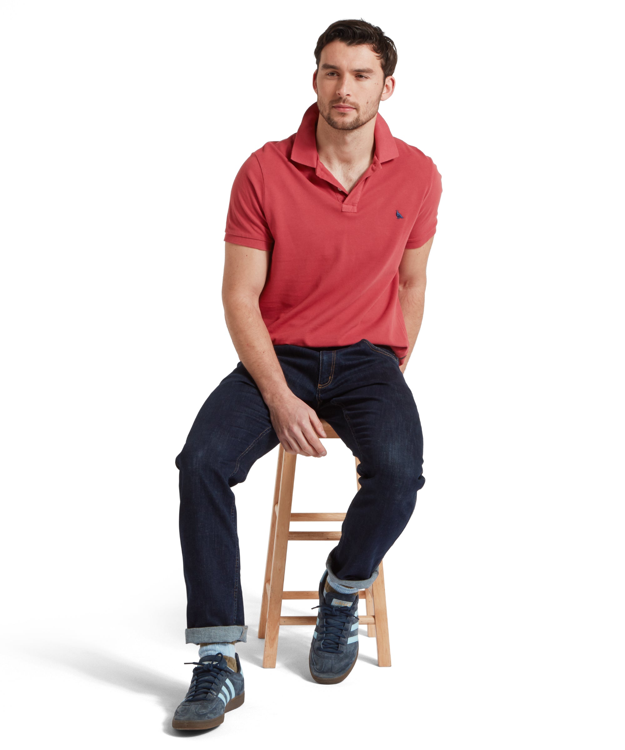 A full height mage of a male model wearing a red Schöffel polo shirt with a small embroidered bird logo on the chest, paired with dark denim jeans, a tan leather belt and navy trainers. He is sitting in a relaxed pose on a wooden stool.