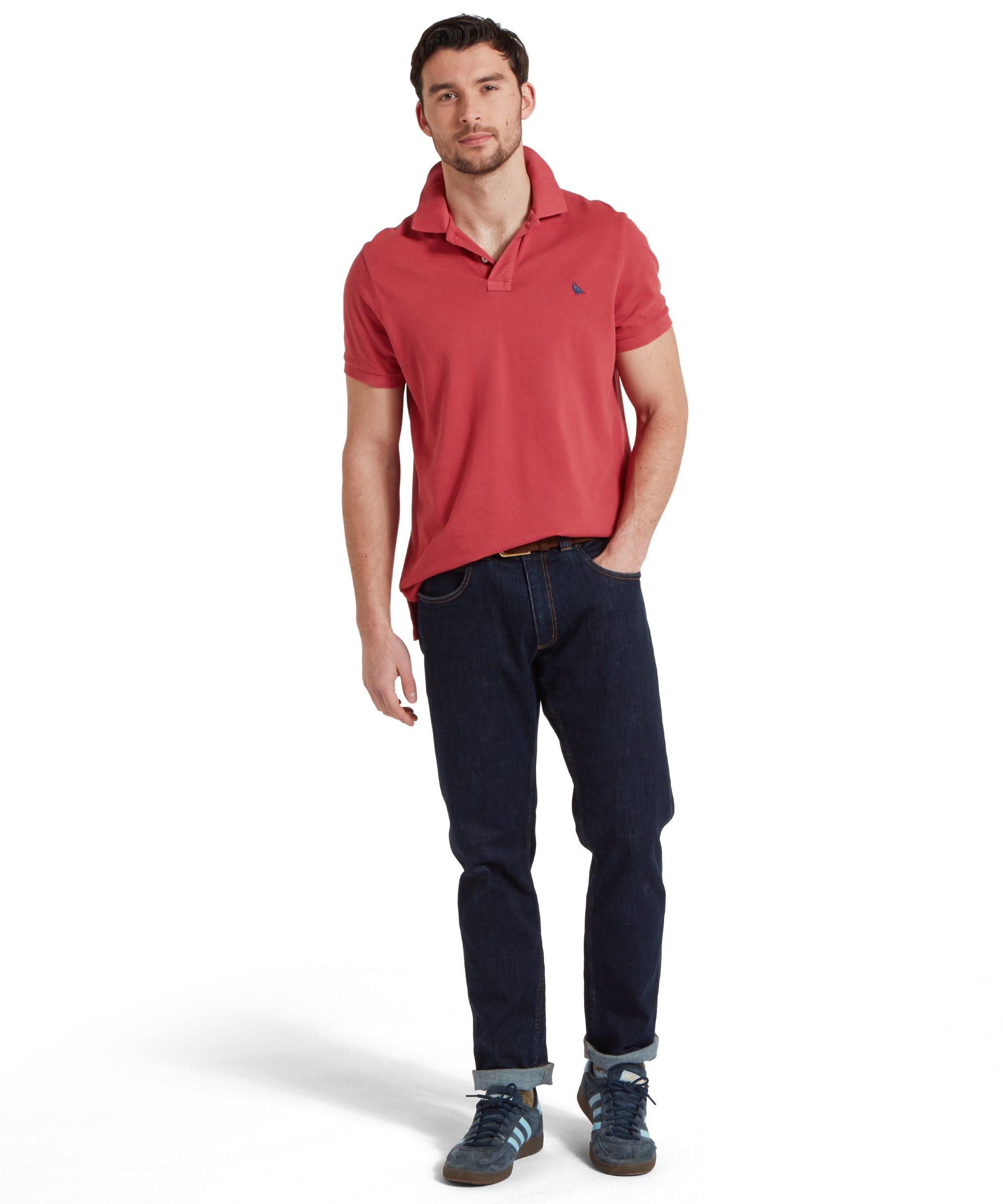 A full height image of a male model wearing a red Schöffel polo shirt with a small embroidered bird logo on the chest, paired with dark denim jeans, a tan leather belt and navy trainers. He is standing in a relaxed pose with one hand in his pocket.