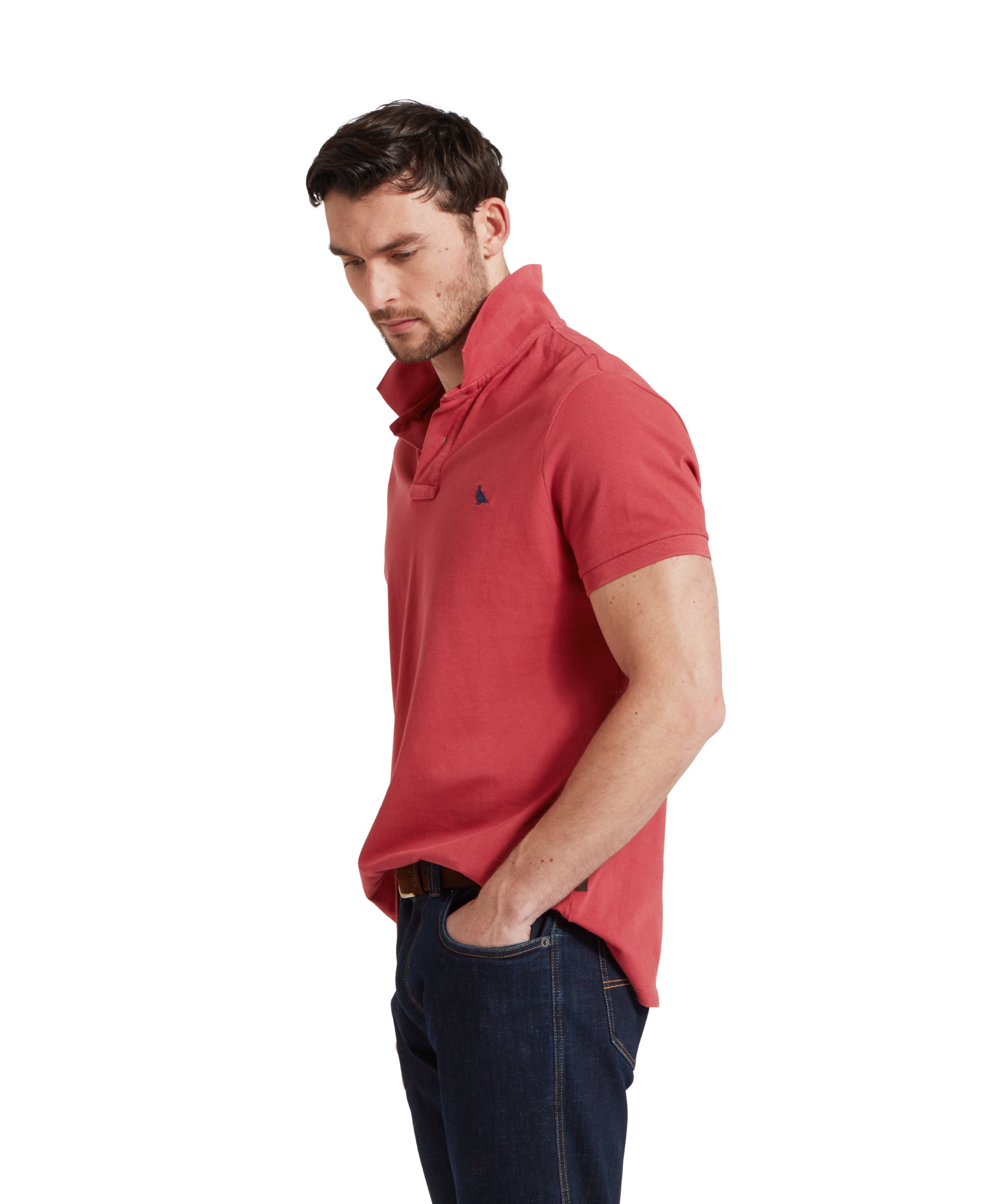 A side view image of a male model wearing a red Schöffel polo shirt with a small embroidered bird logo on the chest, paired with dark denim jeans and a tan leather belt. He is standing in a relaxed pose with one hand in his pocket.