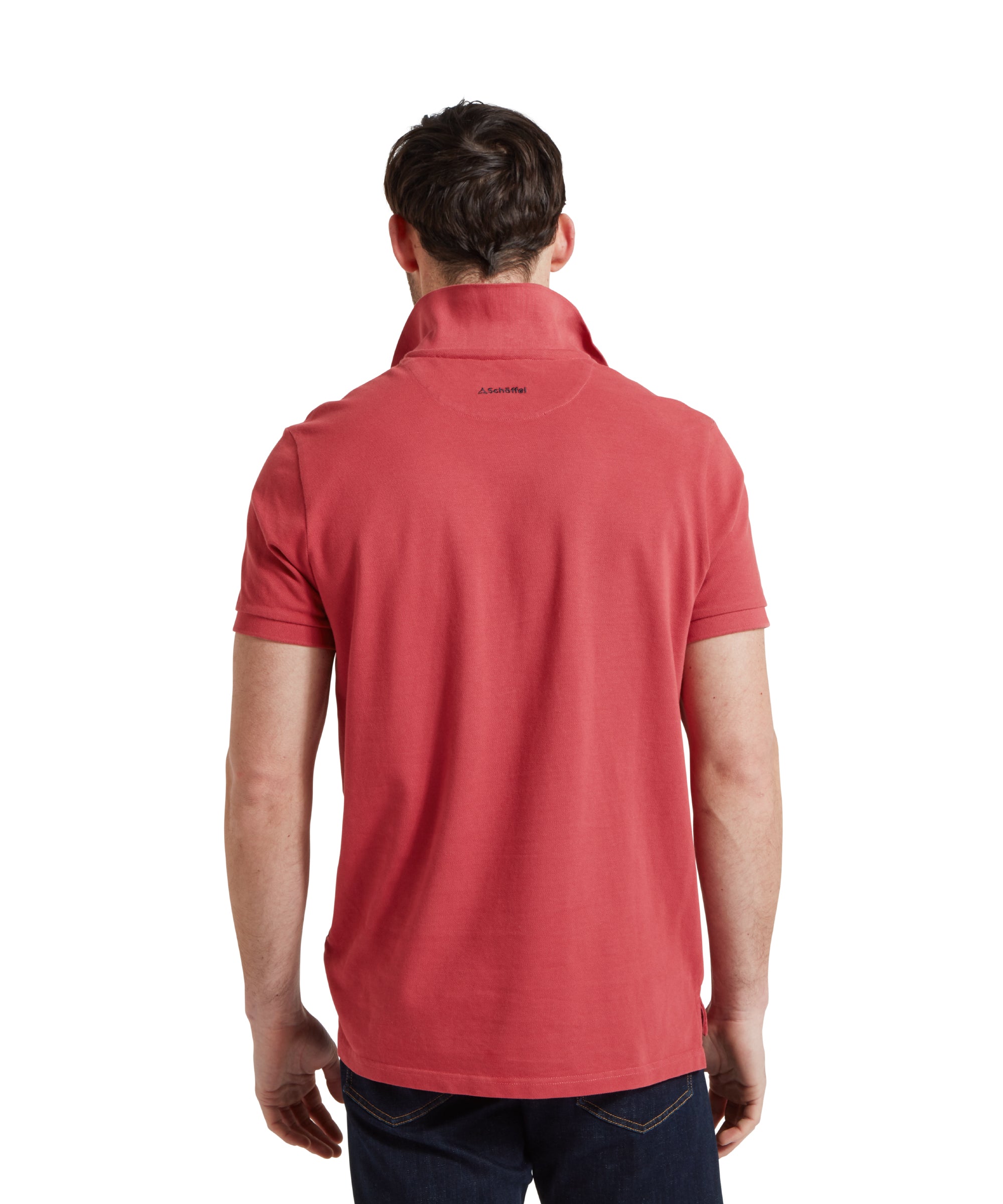 A back view image of a male model wearing a red Schöffel polo shirt with a small embroidered bird logo on the chest, paired with dark denim jeans and a tan leather belt.