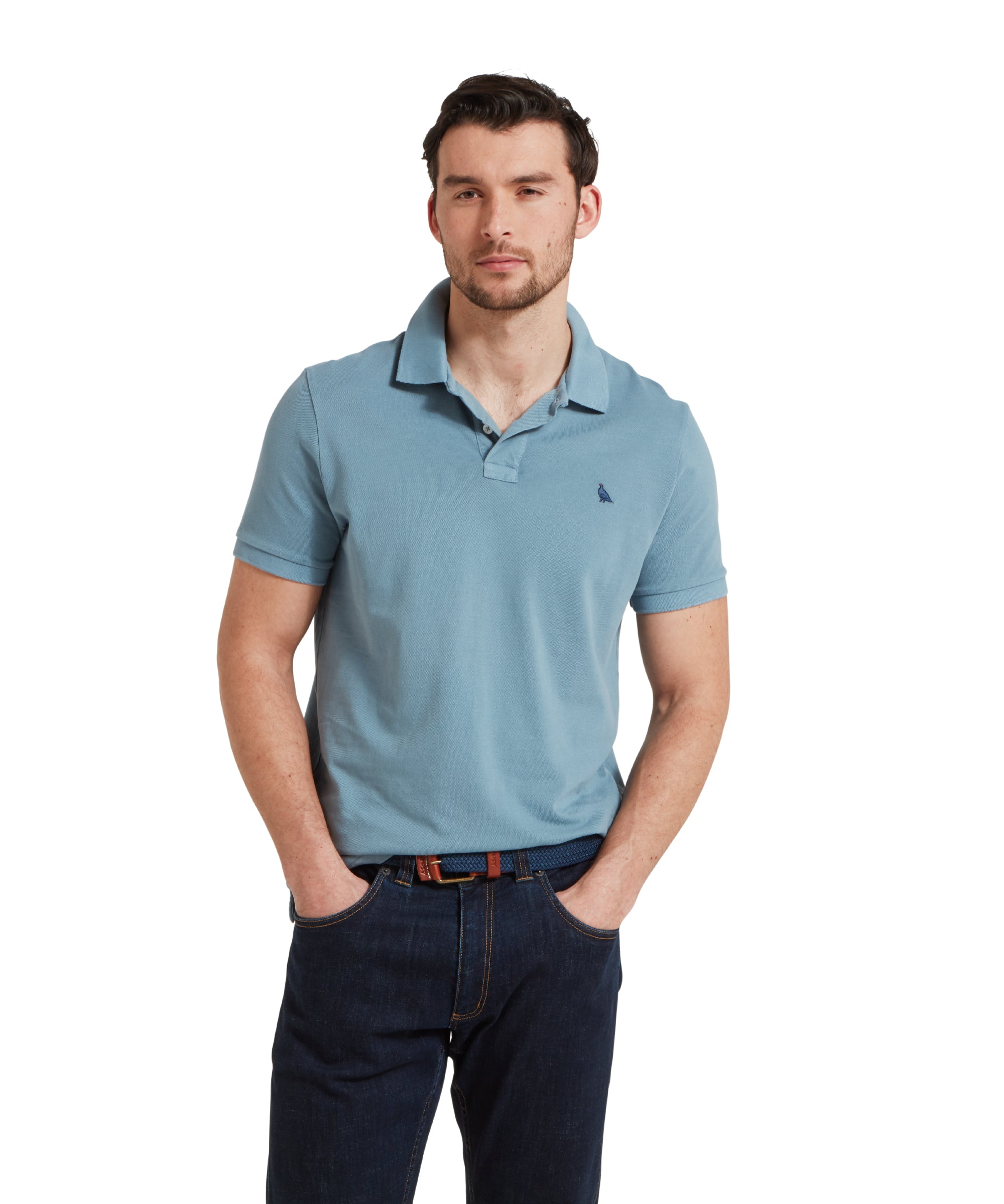 A close up image of a male model wearing a blue Schöffel polo shirt with a small embroidered bird logo on the chest, paired with dark denim jeans and a navy belt. He is standing in a relaxed pose with one hands in his pockets.