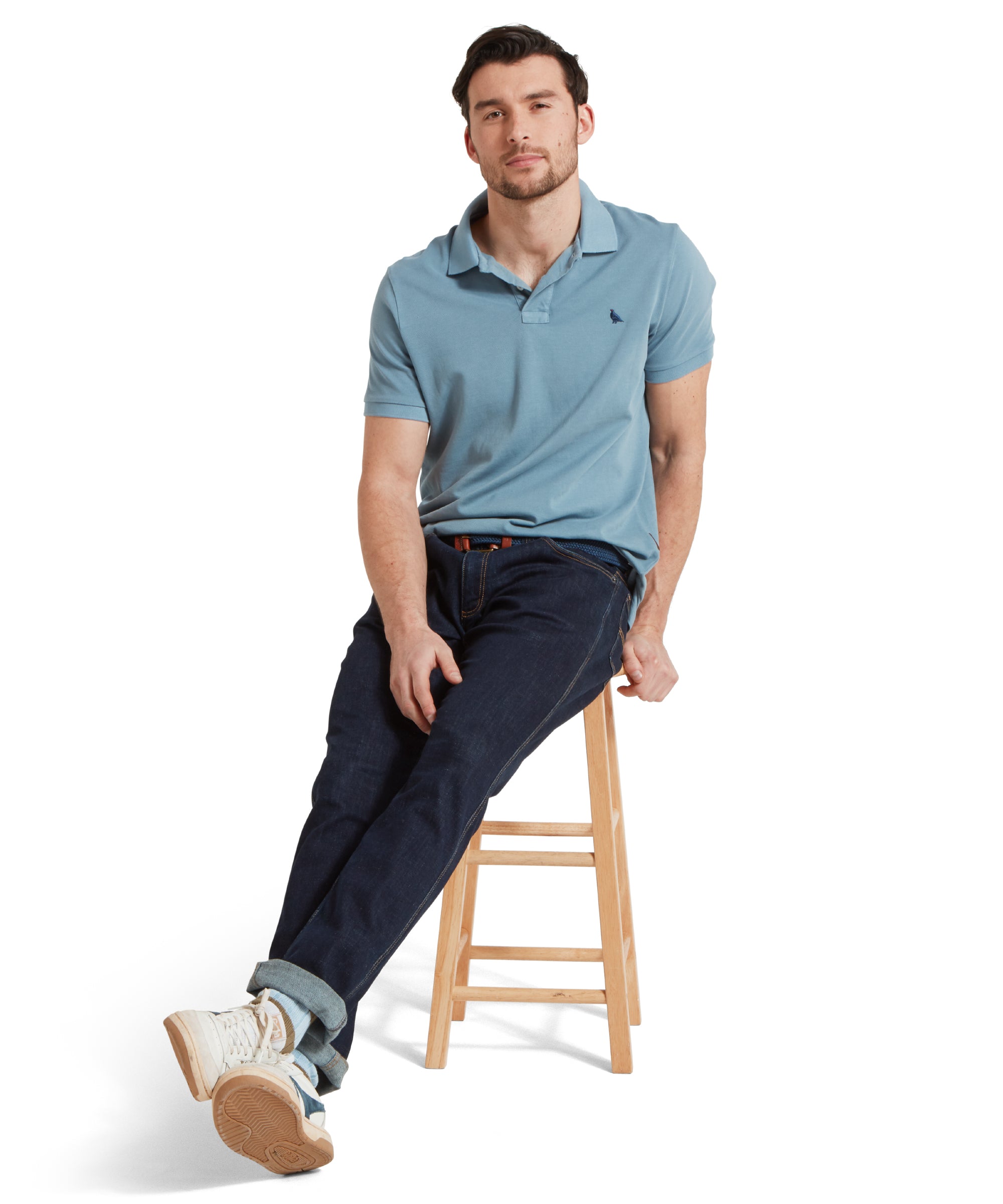 A full height image of a male model wearing a blue Schöffel polo shirt with a small embroidered bird logo on the chest, paired with dark denim jeans, a navy belt and white trainers. He is sitting in a relaxed pose on a wooden stool.