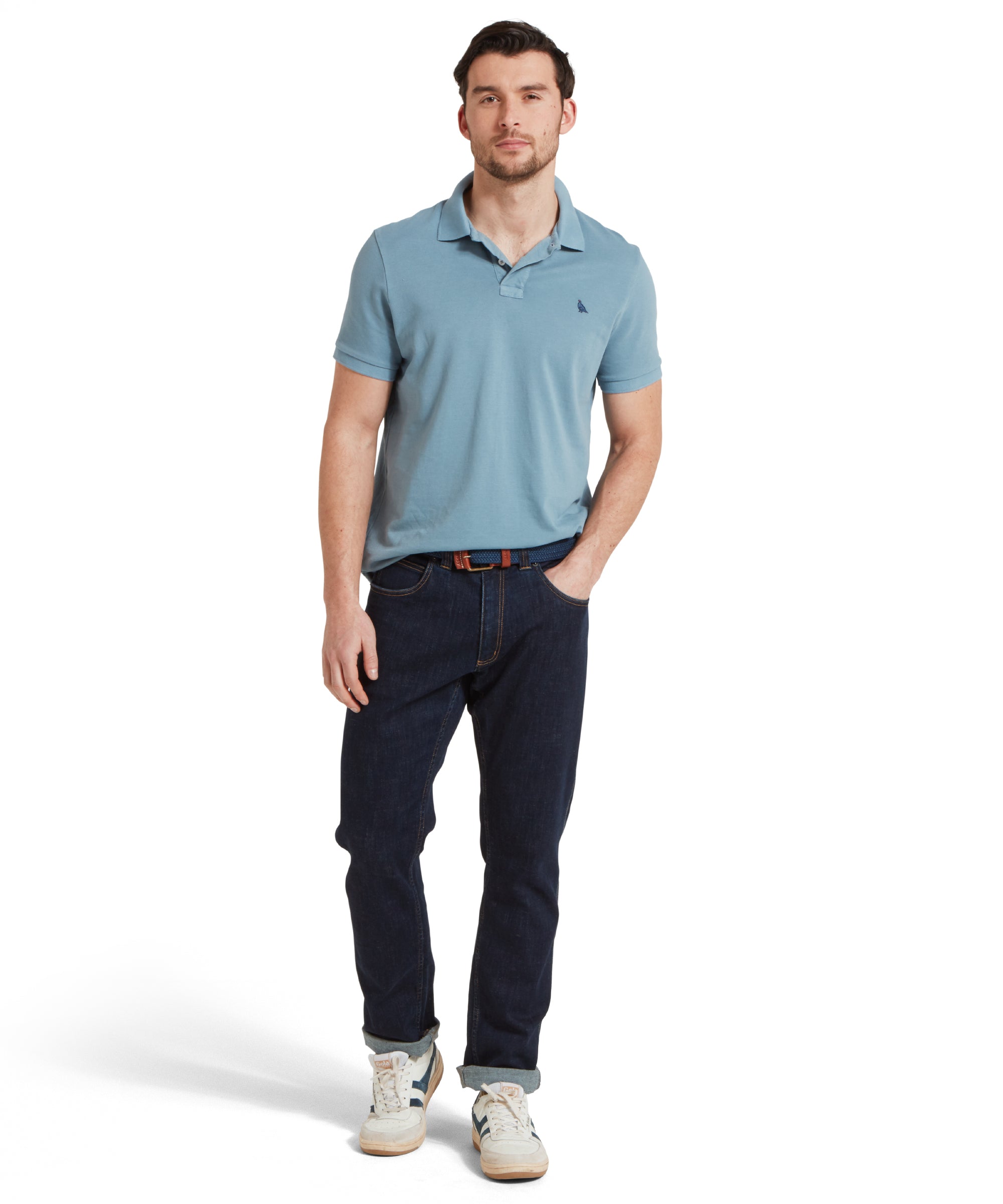 A full height image of a male model wearing a blue Schöffel polo shirt with a small embroidered bird logo on the chest, paired with dark denim jeans, a navy belt and white trainers. He is standing in a relaxed pose with one hand in a pocket.