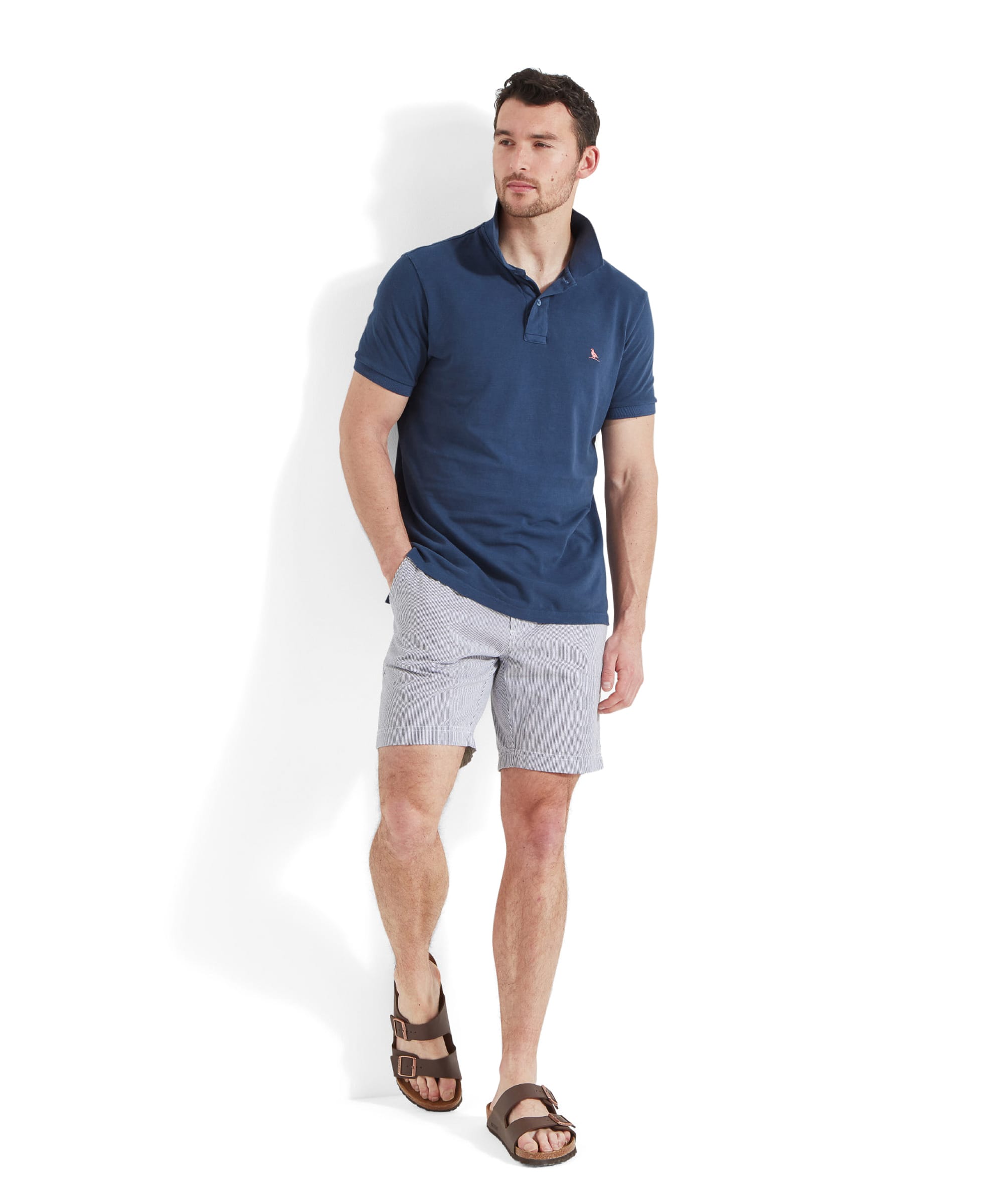 Full body view of a man walking casually in the Schöffel St Ives Garment Dyed Polo Shirt for Men in Blue with striped shorts and brown leather sandals. He's looking to his left with a hand placed in his shorts pocket.