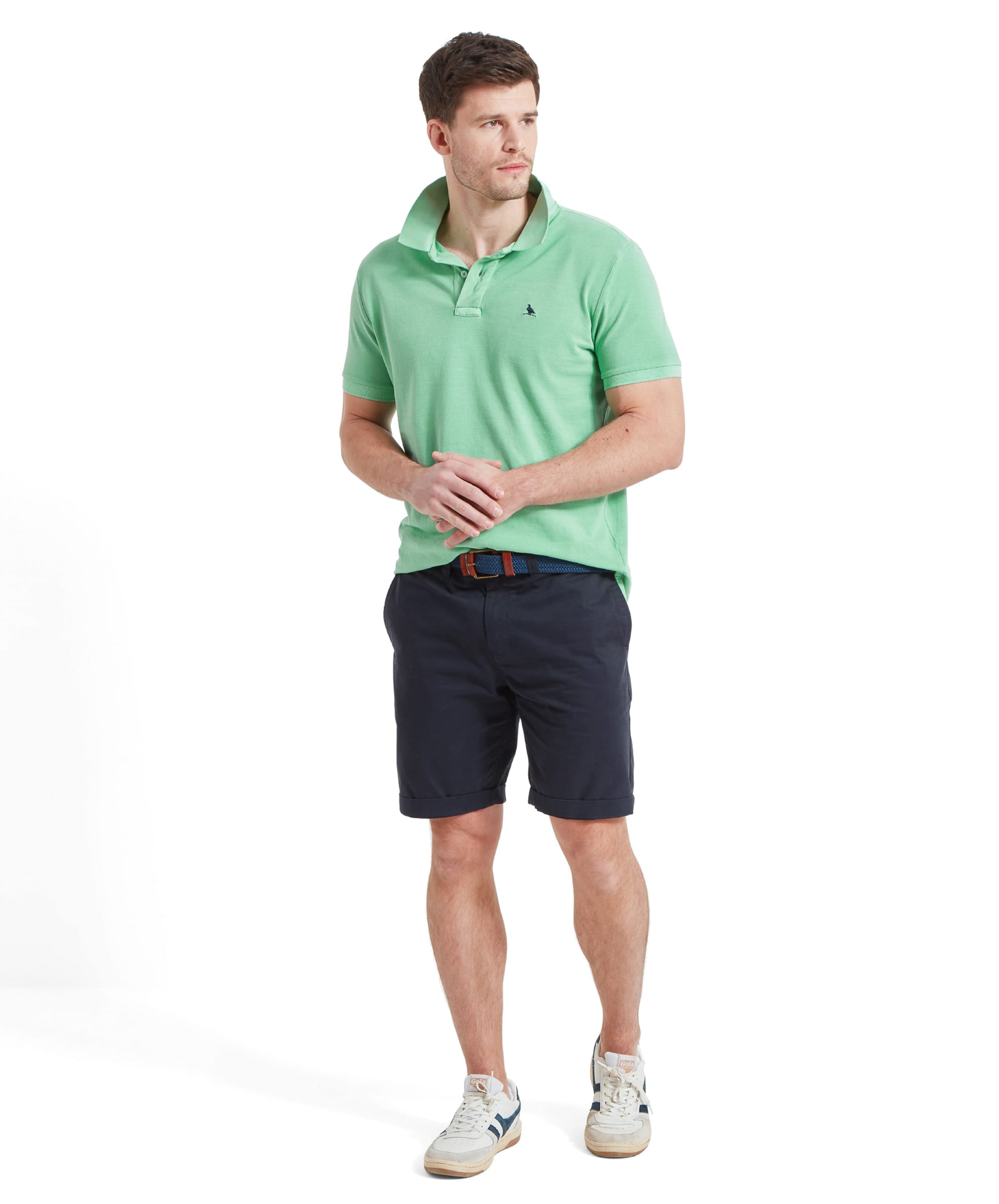 Full view of a man modelling the Schöffel St Ives Garment Dyed Polo Shirt for Men in Green with the collar popped, his hands held together at the front. He wears it with navy shorts and a blue woven belt, and white trainers.