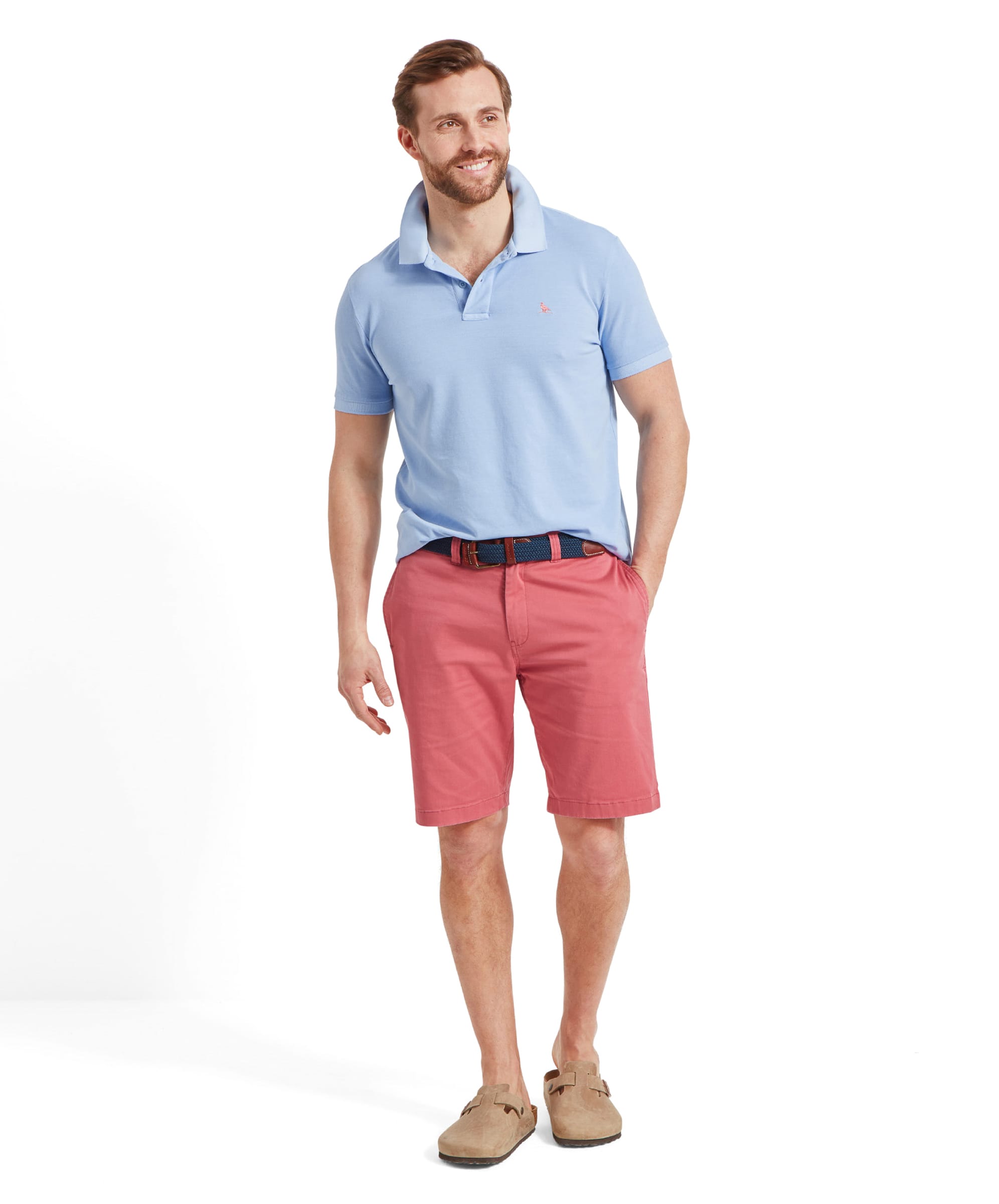 Full view of a man modelling the Schöffel St Ives Garment Dyed Polo Shirt for Men in Light Blue with his right hand in his shorts pocket looking away. He wears the shirt collar popped up, pink chino shorts and a navy woven belt with light beige slip on mules.