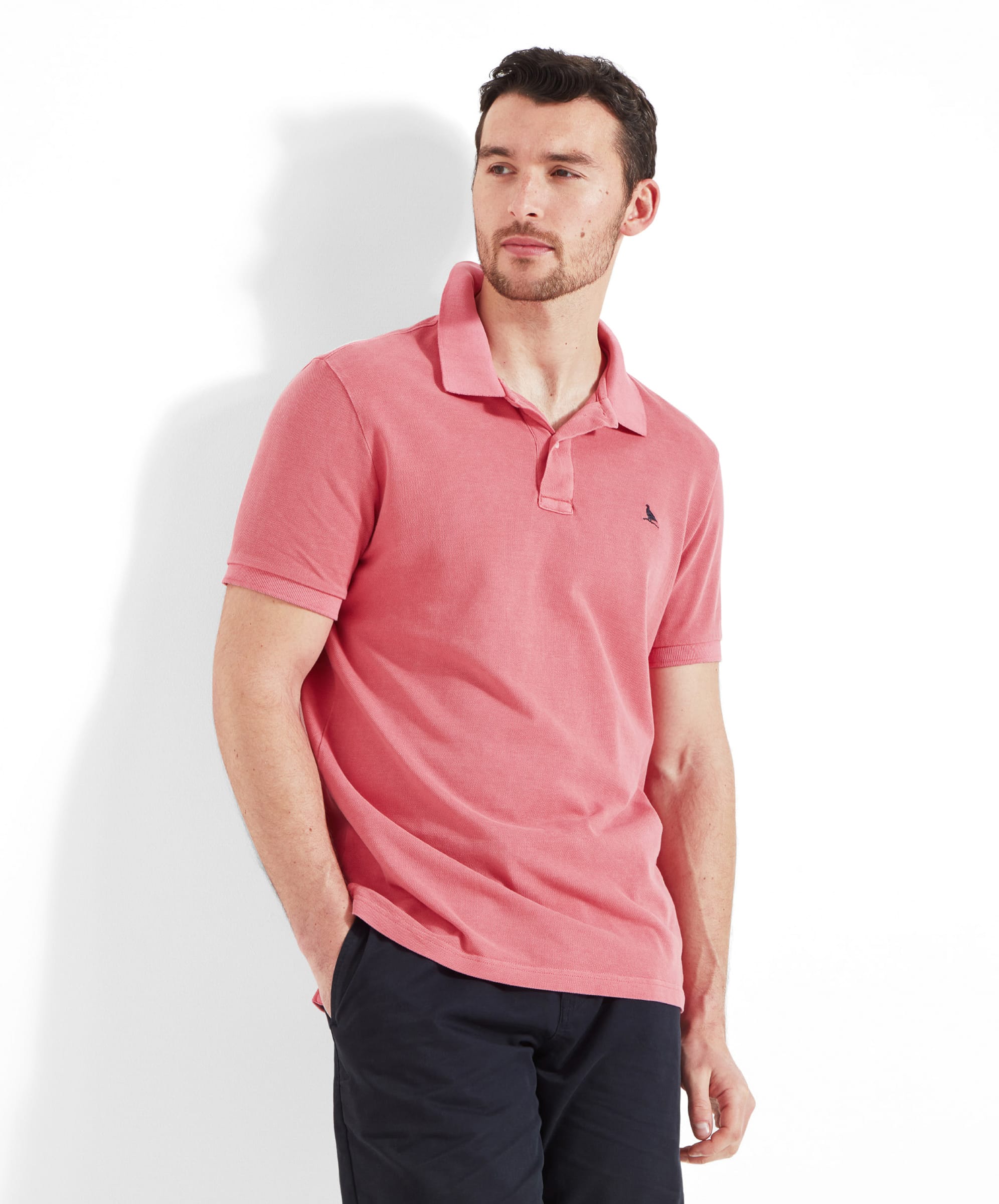 A man wears the Schöffel St Ives Garment Dyed Polo Shirt for Men in Pink with navy chino shorts. He leans casually against a wall with his left hand in his shorts pocket.