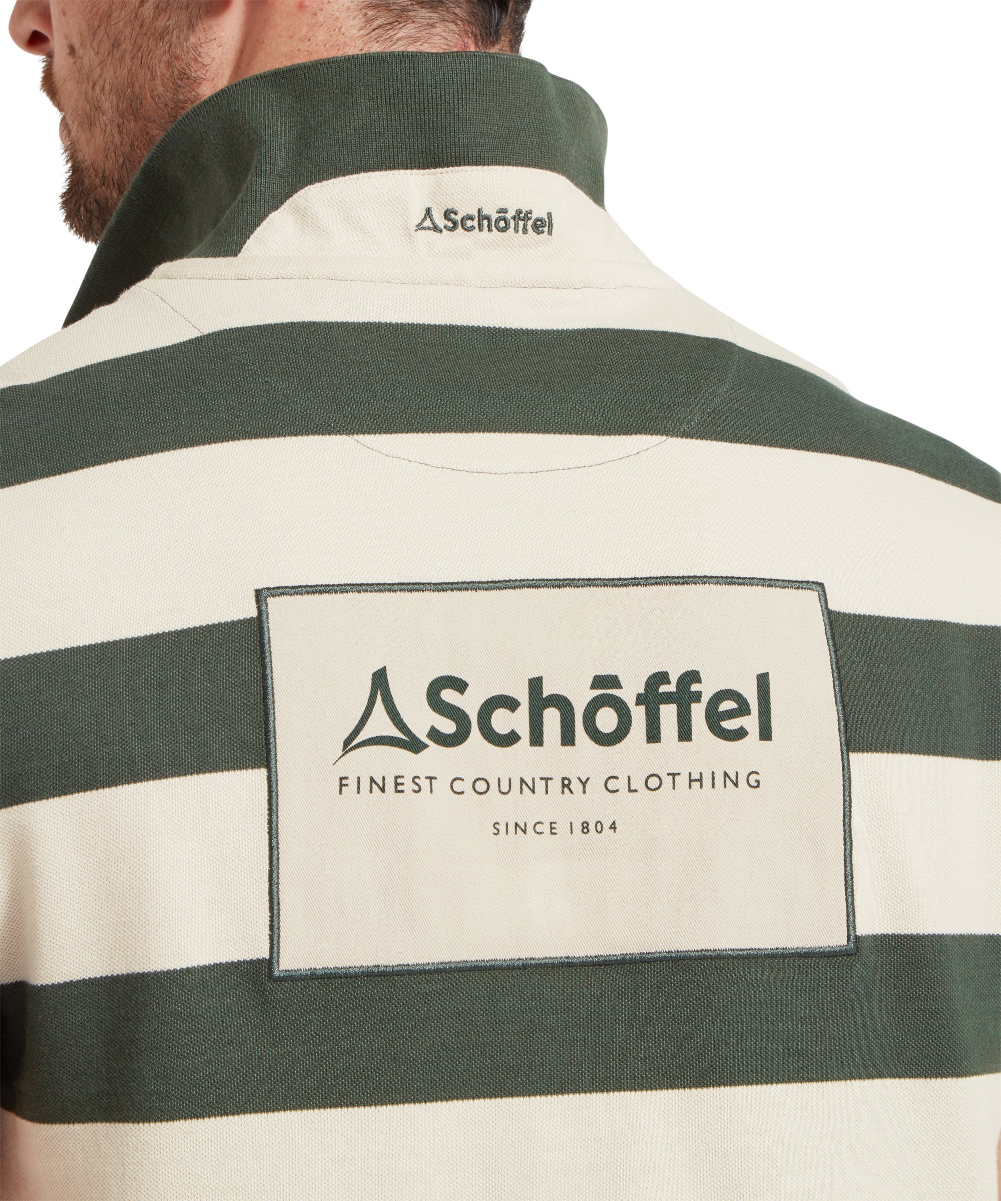 A close up image of a male model wearing a green and beige striped Schöffel polo shirt with a large "Schöffel" logo on the back.