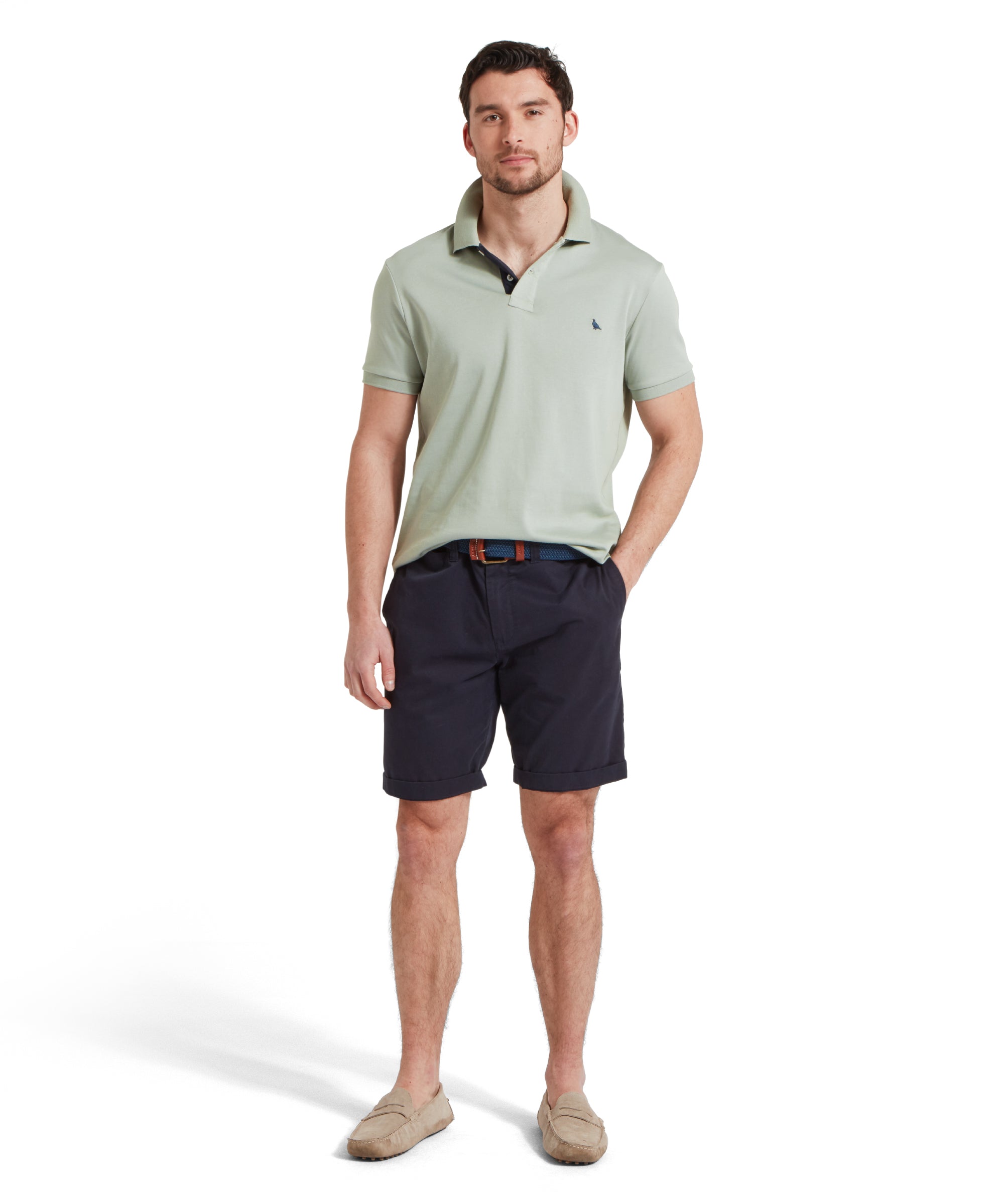 A full-height image of a male model wearing a light green Schöffel t-shirt with a small embroidered bird logo on the chest, paired with navy shorts, a blue woven belt and tan loafers. He is standing in a relaxed pose with one hand in his pocket.