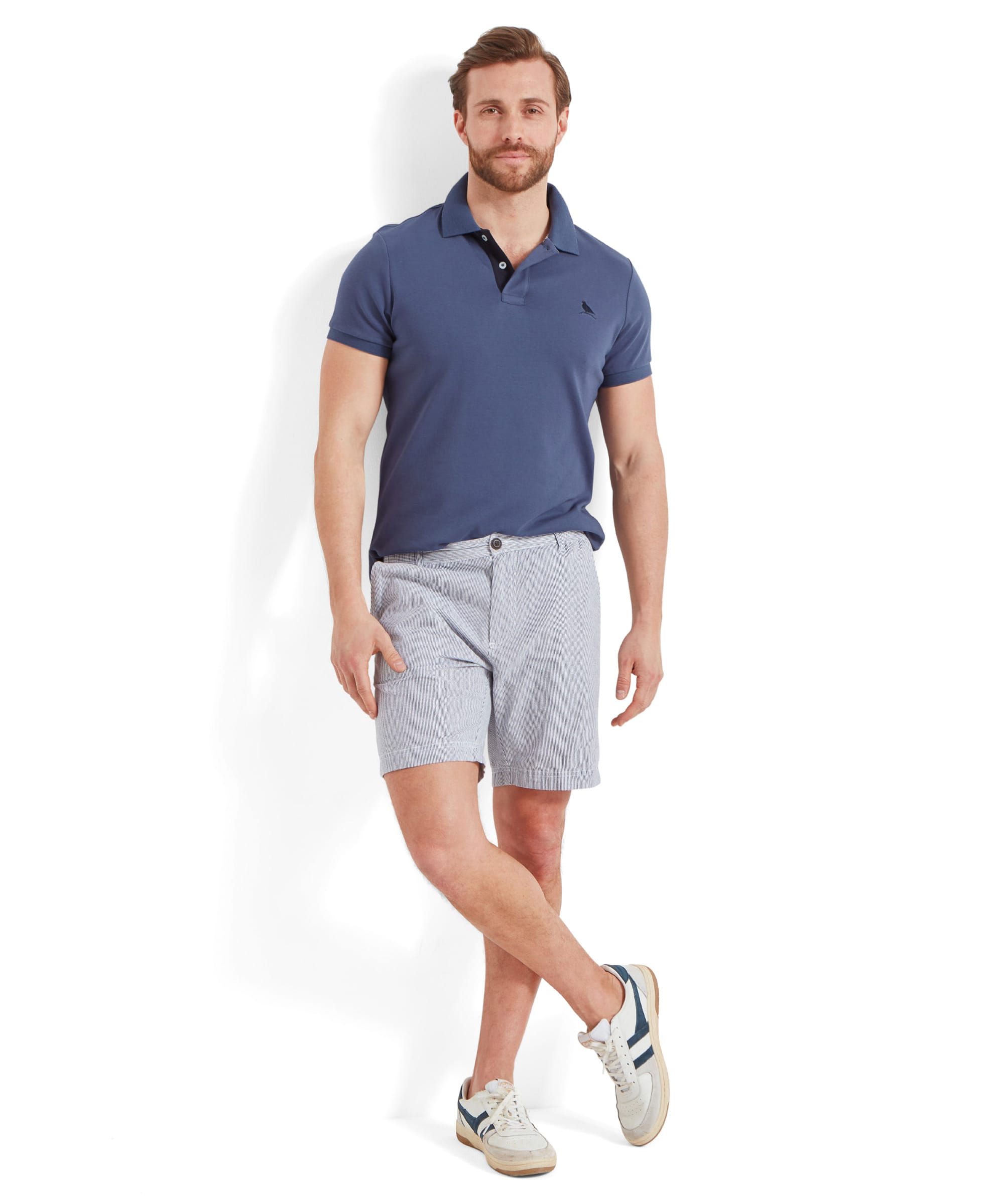 Full-body view of a man wearing the Schöffel St Ives Jersey Polo Shirt for Men in Blue paired with light-colored shorts. He stands casually with one leg crossed over the other.