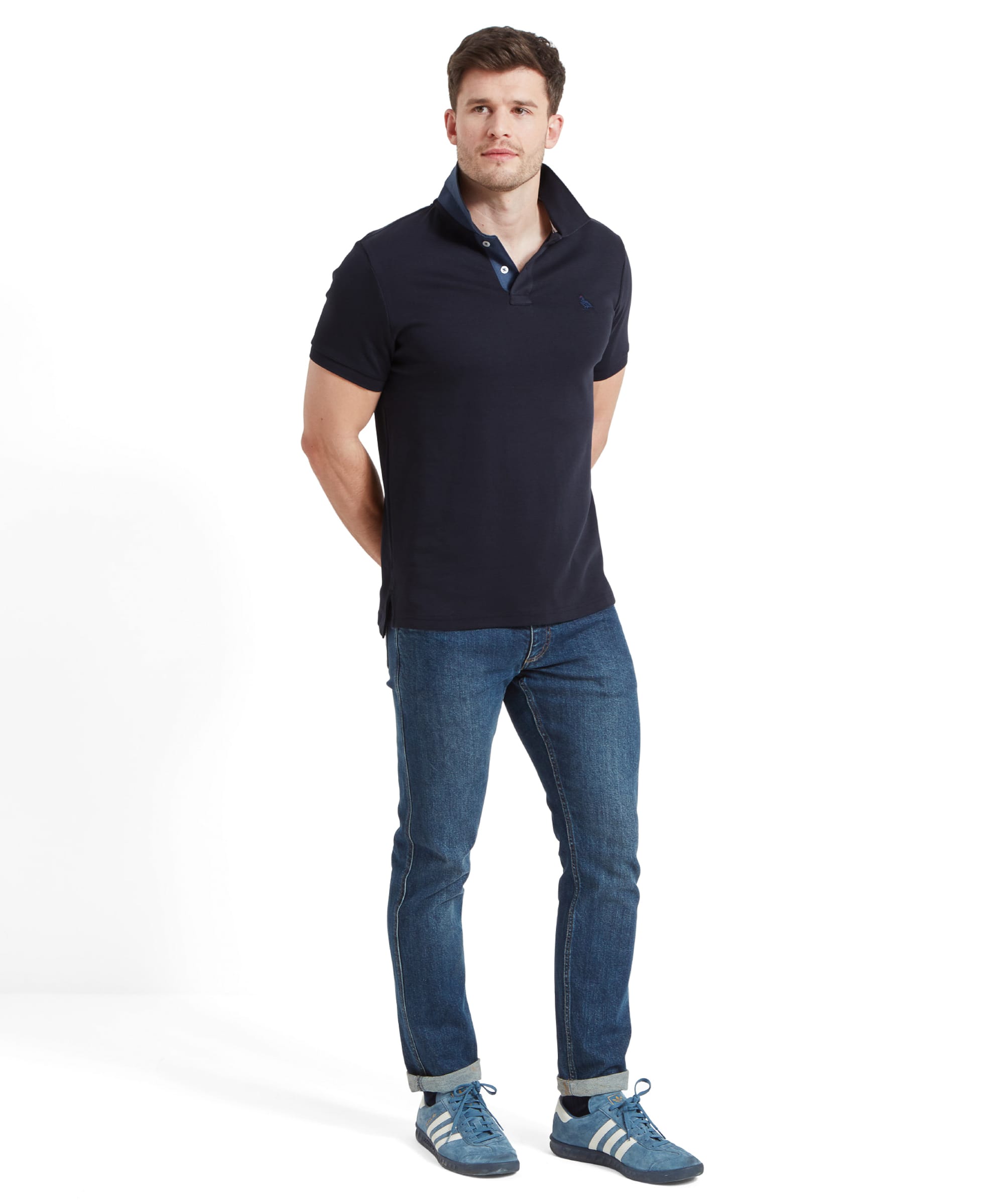 Full-body view of the man wearing the navy polo shirt, standing casually with his hands behind his back. The outfit includes blue trainers, giving a laid-back and casual appearance.