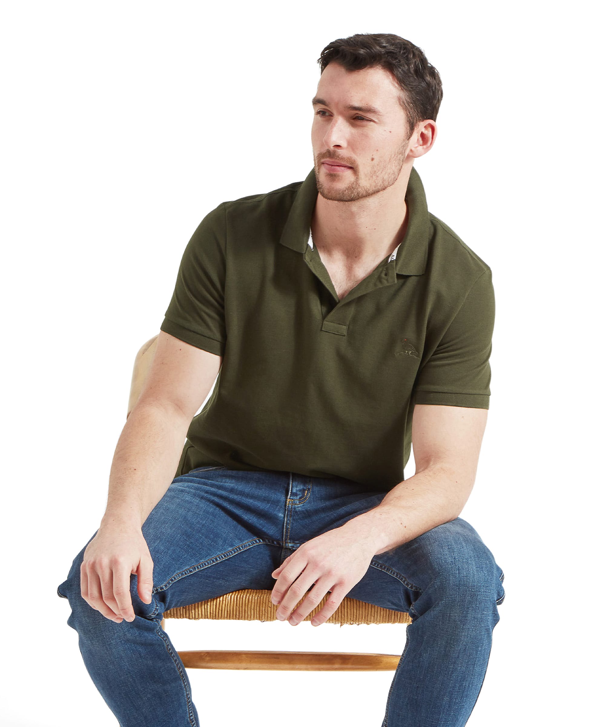 A man wearing the Schöffel Men's St Ives Jersey Polo Shirt in dark green, paired with blue jeans. The shirt features a subtle embroidered bird logo on the left chest and has a classic polo collar. He is seated on a wooden chair, looking off to the side.