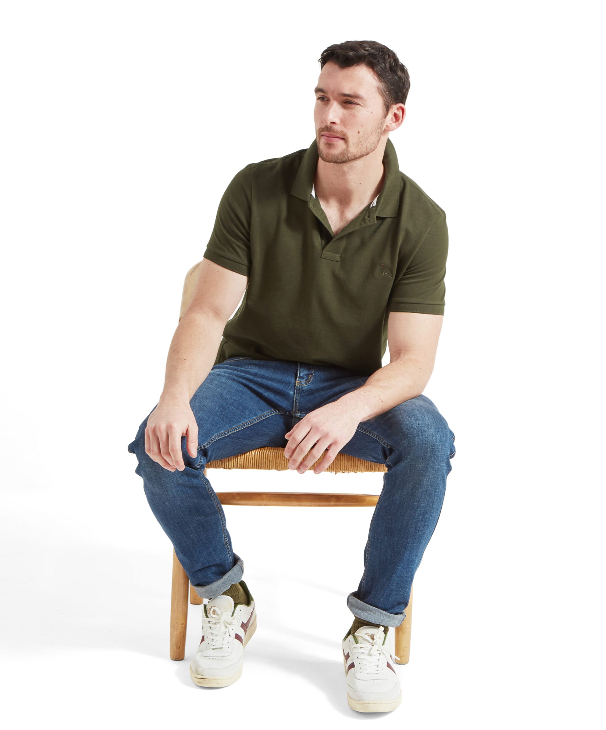 Full-body view of the man sitting casually on a chair, dressed in the dark green polo shirt and blue jeans, with white trainers. The pose and attire give a relaxed, casual vibe.