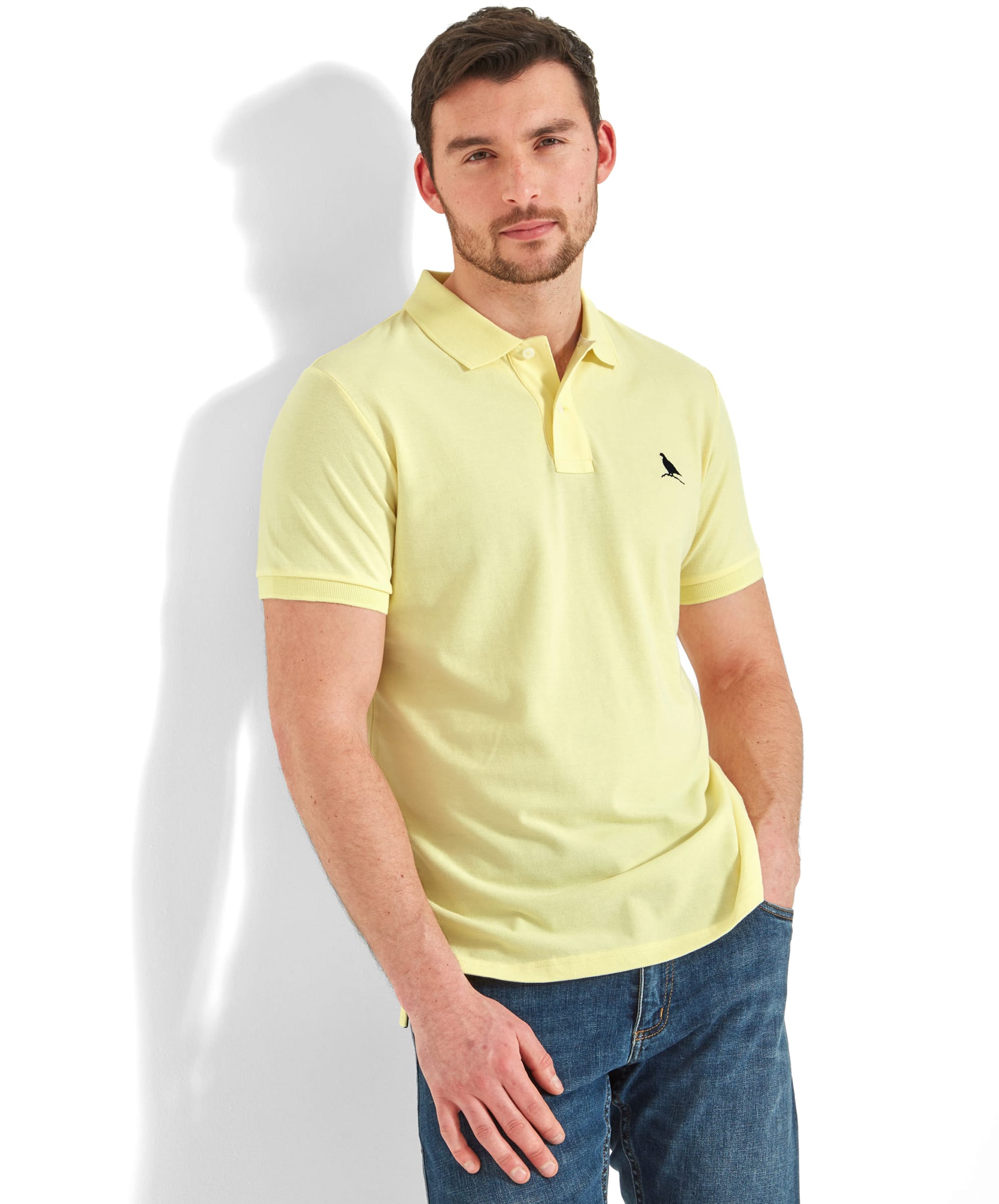 A man wearing the Schöffel St Ives Polo Shirt for Men in Light Yellow, paired with blue jeans. The shirt features a small embroidered bird logo on the left chest and has a classic polo collar. He stands casually against a white wall.