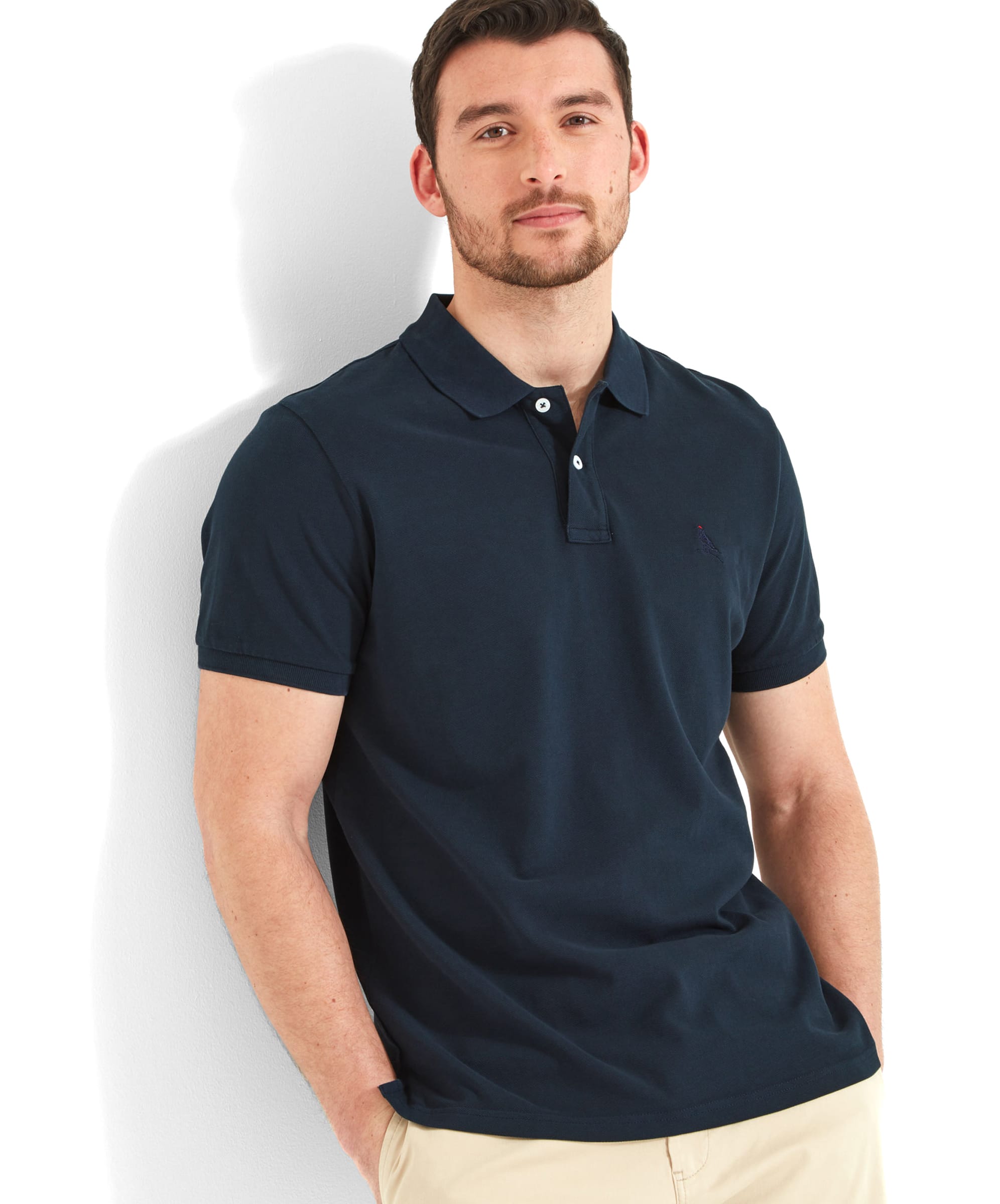 A man wearing the Schöffel Men's St Ives Jersey Polo Shirt in navy, paired with beige shorts. The shirt features a small embroidered Ptarmigan logo on the left chest and has a classic polo collar. He stands casually against a white wall, with a relaxed expression.