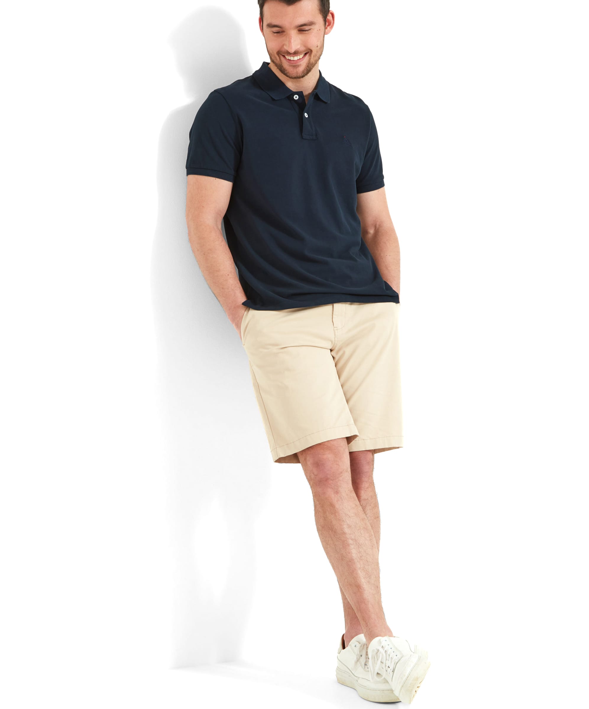 Full-body view of the man wearing the Schöffel St Ives Polo Shirt for Men in Navy, leaning casually against a wall. The outfit includes white trainers, creating a laid-back, comfortable look.