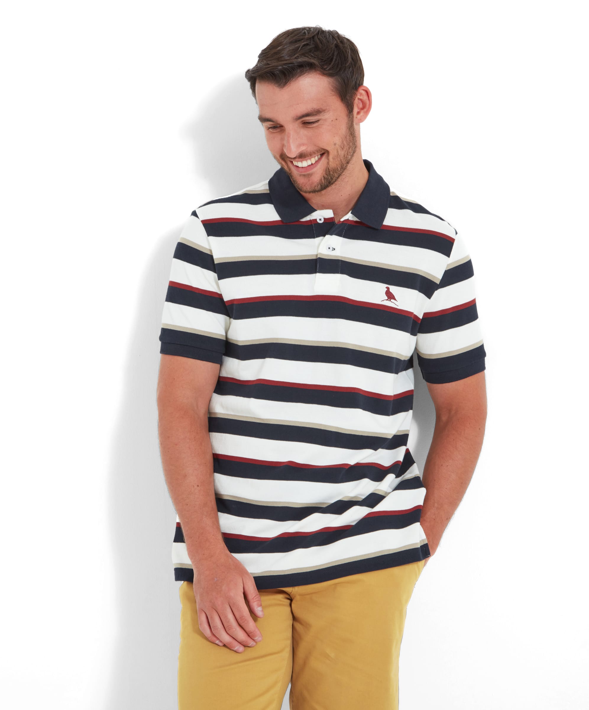 A man wearing the Schöffel St Ives Polo Shirt for Men in Red with horizontal stripes in navy, red, and white. The shirt features a classic polo collar and a small embroidered bird logo on the left chest. The man stands casually against a white wall, smiling.