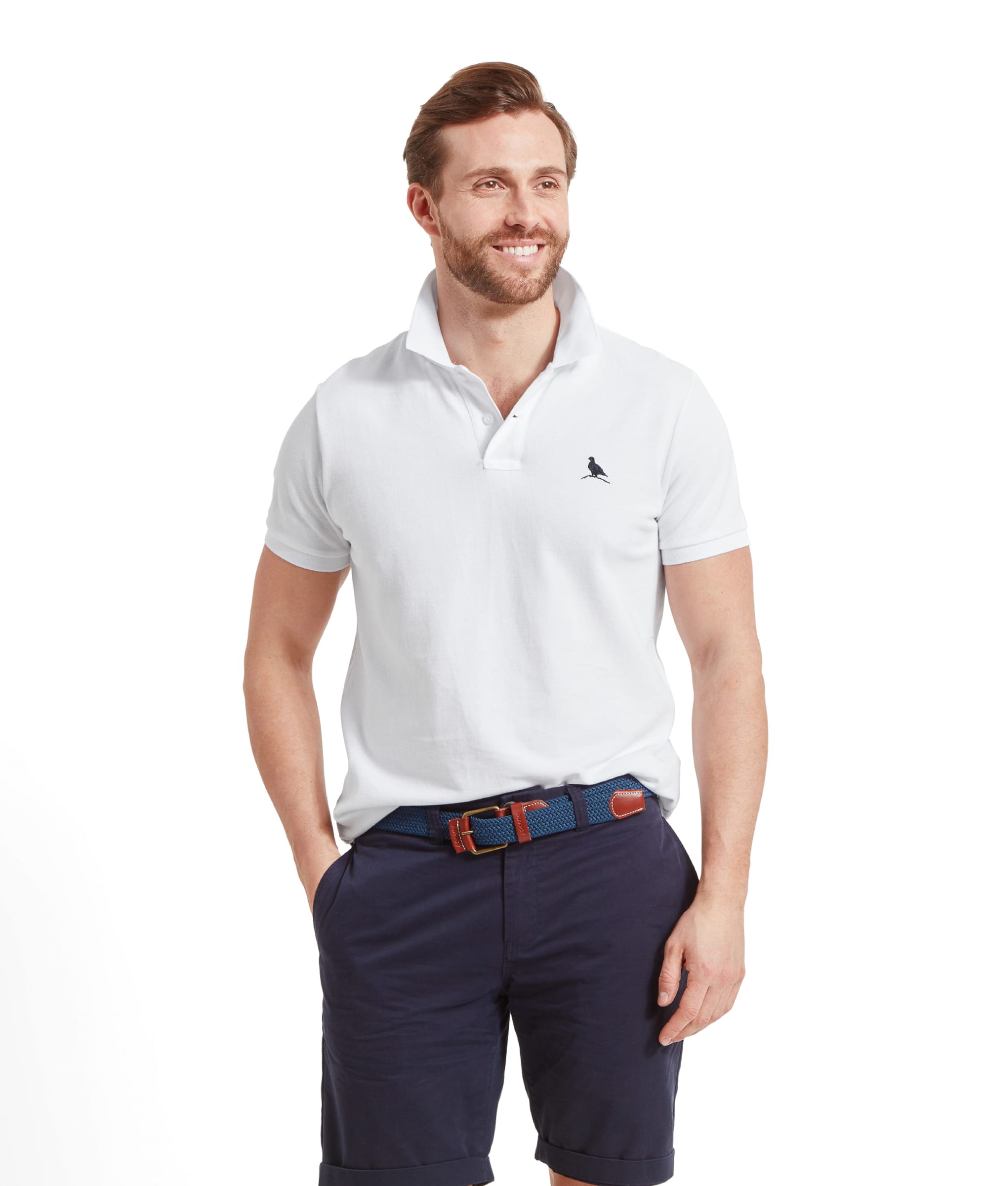 A man wearing a Schöffel St Ives Polo Shirt for Men in White stands smiling with the shirt collar turned up, paired with navy shorts and a blue belt.