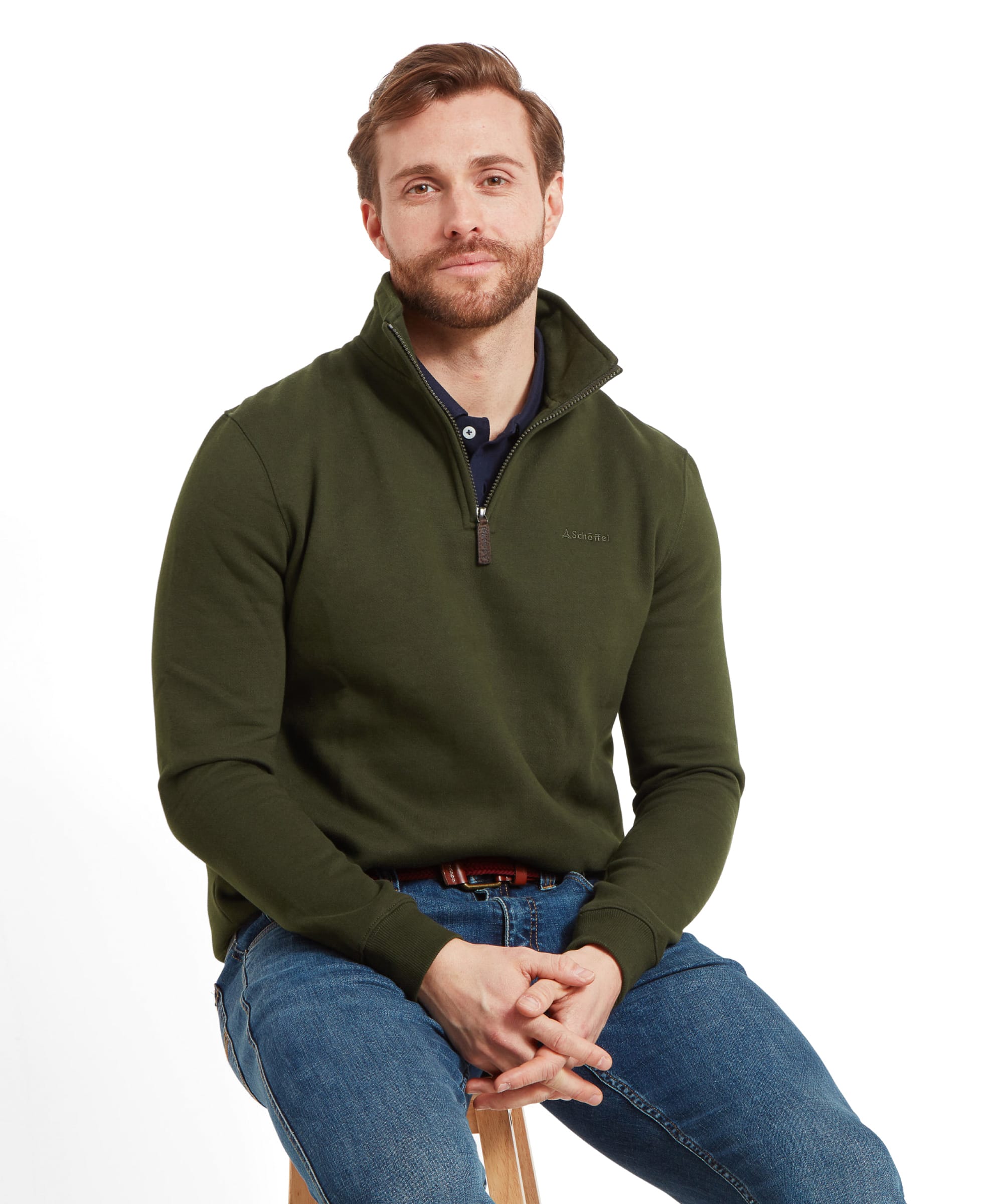 A man sits on a stool, wearing a Schöffel St Merryn Sweatshirt for Men in Dark Green with the Schöffel logo on the chest, paired with blue jeans and green trainers.