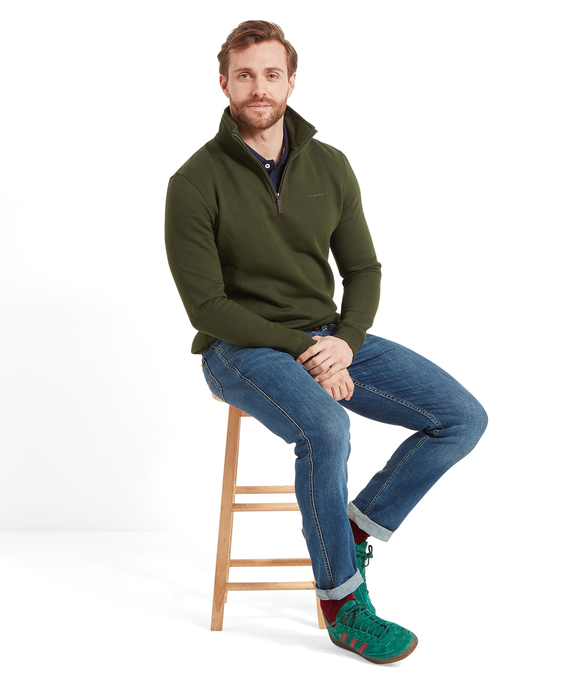 Full body shot of the same man sitting on the stool, showcasing the complete outfit, including the Schöffel St Merryn Sweatshirt for Men in Dark Green, blue jeans, and green trainers with red stripes.