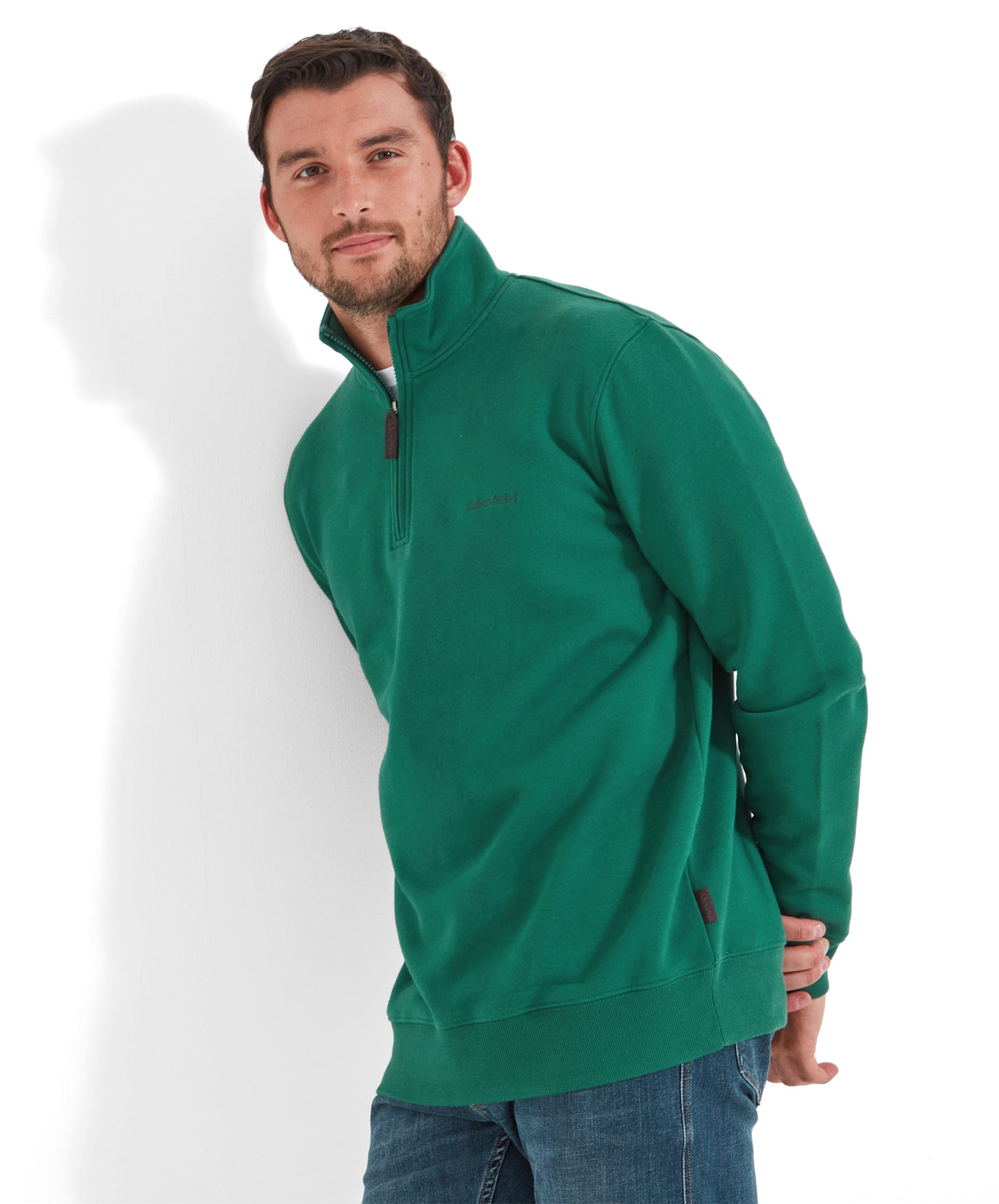 A man wearing a Schöffel St Merryn Sweatshirt for Men in Green, sitting on a wooden stool. He has a short beard and is smiling slightly. The sweatshirt features a small logo on the left chest and is paired with blue jeans and green trainers.