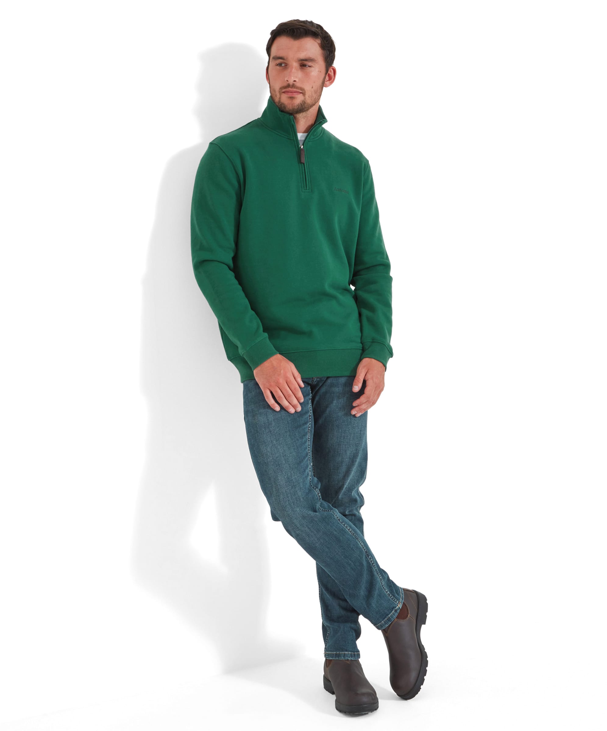 The same man in the Schöffel St Merryn Sweatshirt for Men in Green, sitting casually on the stool with his hands clasped, showing a full view of his outfit including blue jeans and green trainers.